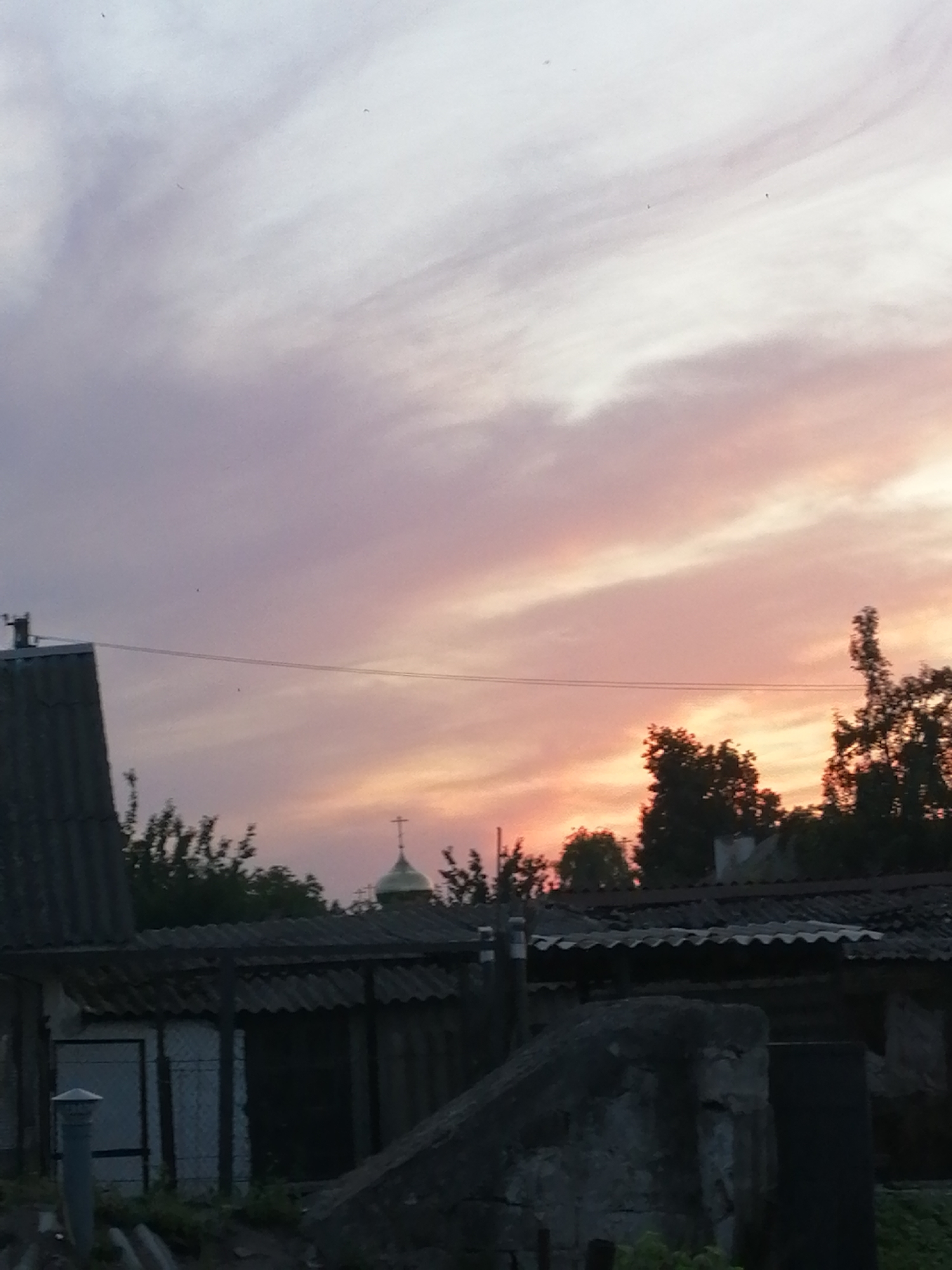 The image depicts a dusky sky with hues of pink, purple, and white, characteristic of sunset or sunrise. In the foreground, there are several low-lying buildings with corrugated roofs, some appearing aged or in disrepair. A golden dome with a cross, likely belonging to a church or chapel, is visible above the rooftops, silhouetted against the colorful sky. Trees with dark foliage are scattered throughout the scene, some obscuring parts of the buildings. A power line stretches horizontally across the frame. The overall impression is of a rural or semi-rural setting, possibly within the city of Lanivtsi, Ukraine, given the architectural style and the presence of a religious structure. The scene appears quiet and still, with no people or overt activity visible.