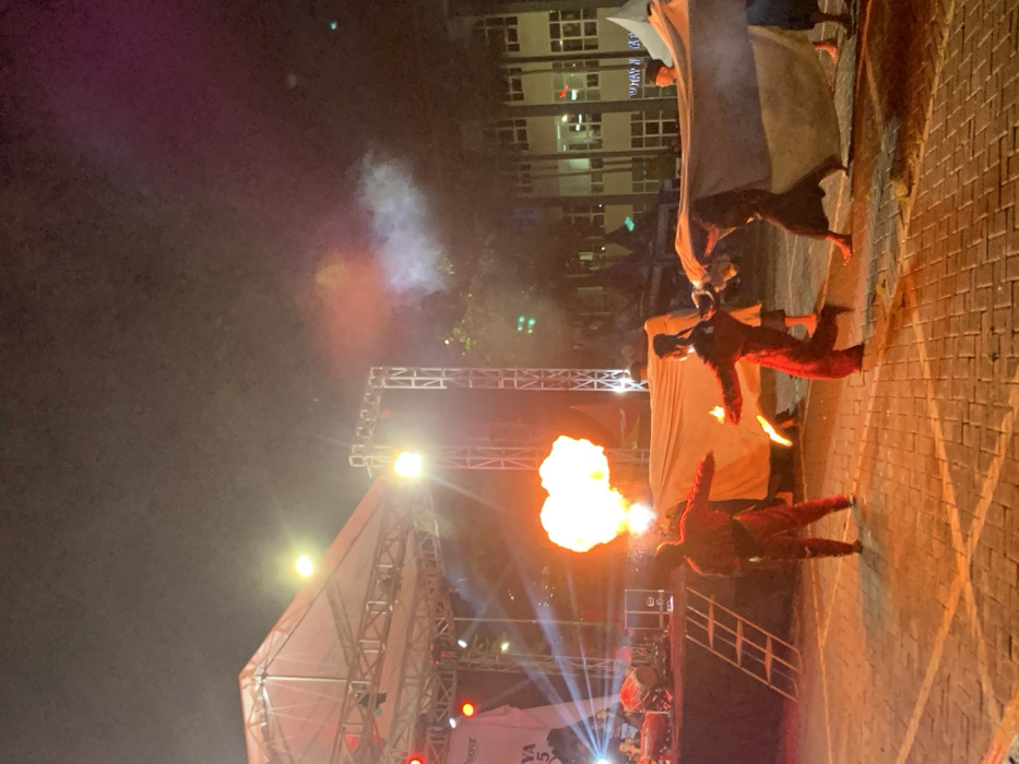 The scene takes place at night in front of a stage in the city of Kecamatan Batu, Indonesia, at the coordinates -7.867 latitude and 112.514 longitude. Five men wearing dark, red, or blue clothing are performing a fire dance in front of a large white sheet. There are red and blue lights illuminating the scene, and a fire is burning, creating a large orange flame. The men are moving their bodies in a coordinated fashion as they maneuver around the fire. The ground is made of brick pavers. There is a ladder on the left side of the scene, which is used for access to the stage, where a large white sheet is suspended. There are buildings in the background, which are lit up. The image appears to have been taken at a festival or other event.