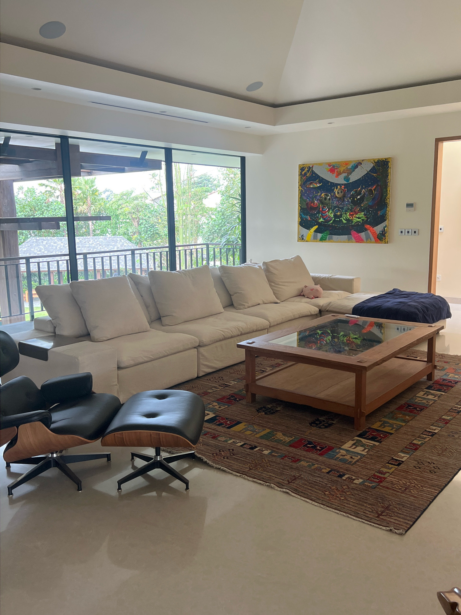 This is a well-lit, interior shot of a modern living room in Ngaglik, Indonesia. The room features a large, light-colored sectional sofa with numerous cushions, positioned near a floor-to-ceiling window that offers a view of lush greenery and an outdoor structure with a tiled roof. In front of the sofa sits a wooden coffee table with a glass top, resting on an intricately patterned rug. To the left of the sofa, two black leather armchairs with wooden accents and matching ottomans are arranged. A colorful, abstract painting hangs on the off-white wall above the sofa. A dark blue throw blanket is placed on the right side of the sectional. The flooring is a smooth, light-colored material, possibly concrete or polished stone. The overall aesthetic is contemporary and comfortable, with a focus on natural light and outdoor views.