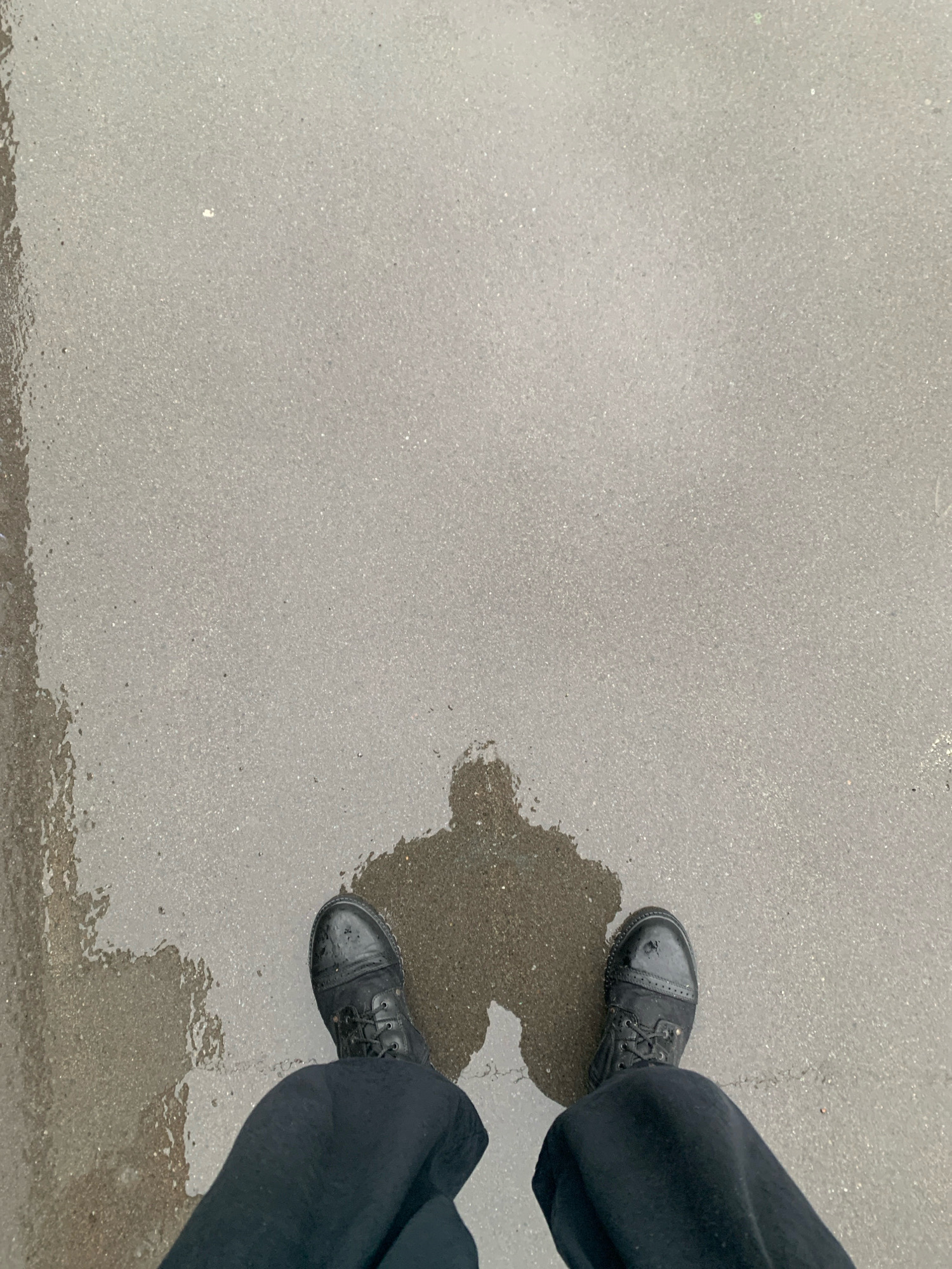 The image shows a pair of black boots standing on a wet, gray asphalt surface. A shadow of the person wearing the boots is cast on the ground. The scene appears to be outdoors, likely in a city, given the asphalt and the general appearance of the environment. The lighting suggests it is daytime, possibly overcast or after rain due to the wetness. There are no other people or identifiable objects in the immediate vicinity. The lower parts of black pants are visible above the boots. The specific location in Arkhangelsk, Russia, cannot be determined from the visual information alone, as there are no distinct landmarks or identifying features.