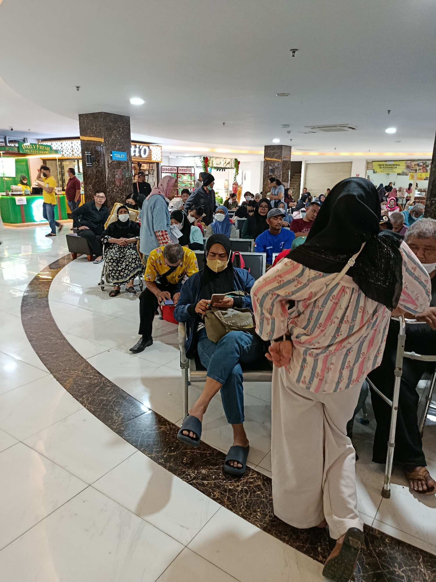 In a brightly lit indoor area with a checkered marble floor, a crowd of people are waiting, some seated on benches, others standing. The environment appears to be a public space, possibly a clinic or waiting area, given the presence of multiple individuals and seating arrangements. In the background, signs indicate commercial establishments, including one for "DAILY FRESH THE FOOD HOUSE" and another with the word "TOILET" visible. The people in the image are diverse in age and attire, with many wearing masks. Some individuals are engaged with their mobile phones, while others appear to be conversing or simply waiting. The scene captures a moment of everyday life in East Jakarta, Indonesia, likely at a busy public facility.