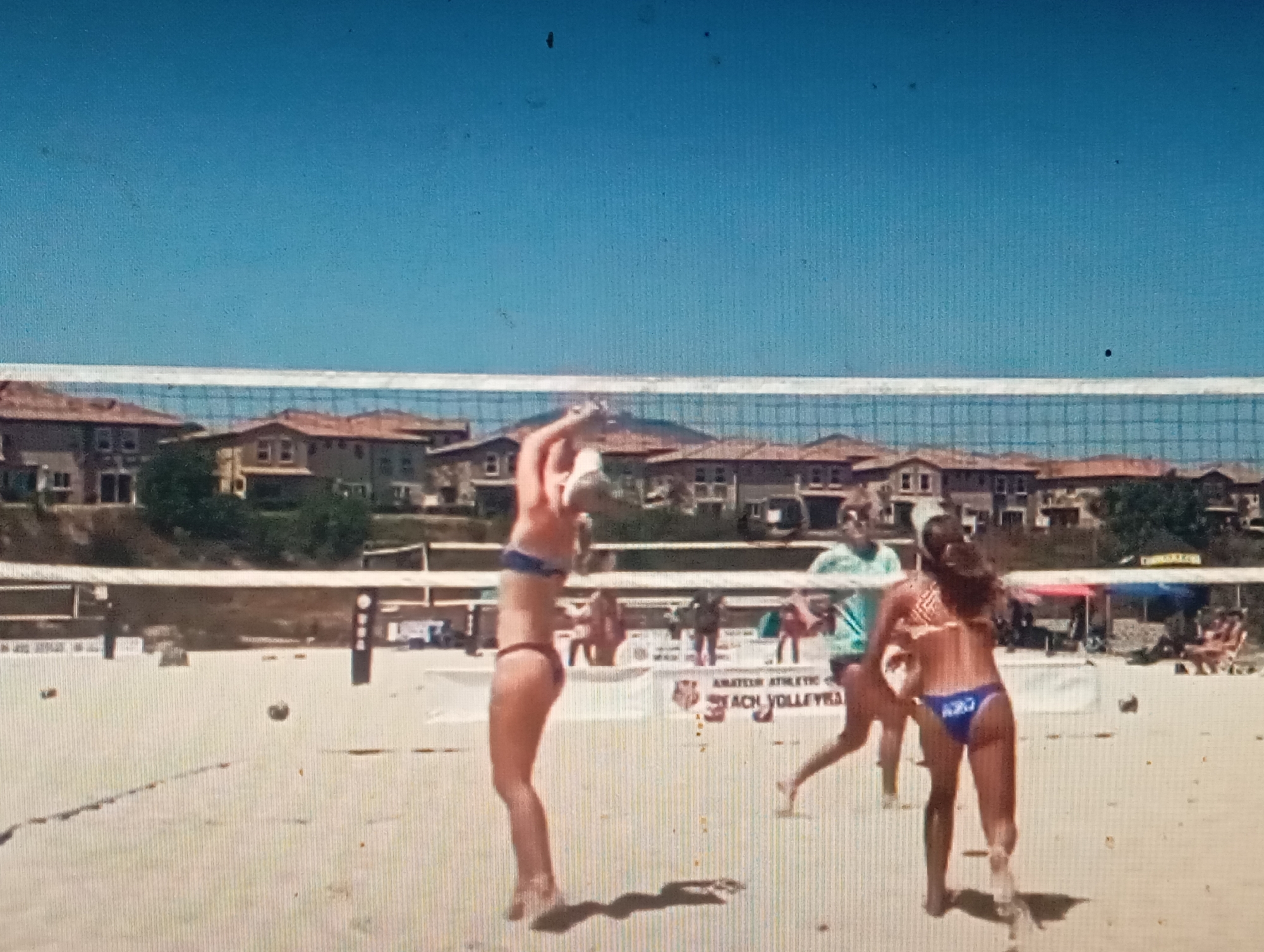 This is a photograph of a beach volleyball game played on a sandy court under a clear blue sky. In the foreground, two women in bikinis are actively playing. The woman on the left is in mid-air, with her left arm raised as if to serve or hit the ball. The woman on the right is facing away from the camera, her left leg extended as she moves. A volleyball net is stretched across the court, separating the players. In the background, there are buildings with terracotta roofs and trees, suggesting a developed area adjacent to the beach. Several spectators are visible in the distance, some sitting in chairs. A banner behind the players reads "AMATEUR ATHLETIC BEACH VOLLEYBALL." The image does not contain any information that would indicate the location is in Sofiivska Borshchahivka, Ukraine.