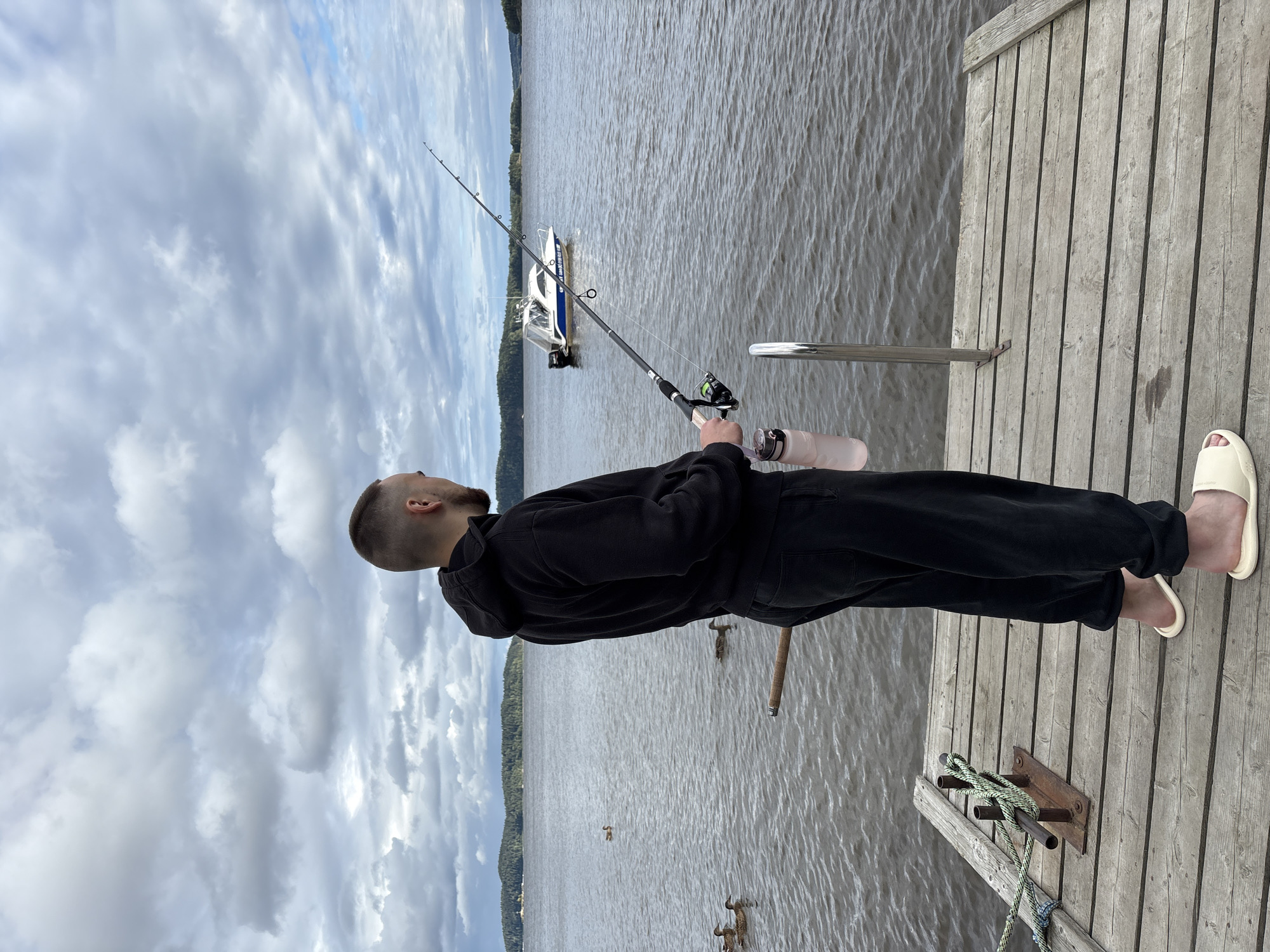 A young man stands on a wooden dock in Rautalakhti, Russia, holding a fishing rod and looking out at the water. He is wearing a black hoodie and black sweatpants, and he has on light-colored slip-on shoes. In the background, a white boat with a blue stripe is visible on the lake. Several ducks are swimming in the water near the dock. The sky is cloudy, and the overall impression is one of a calm, overcast day. The dock is made of weathered wood planks, and a metal railing is attached to its side. A coiled rope is tied to a metal fixture on the dock. The water is choppy, with small waves. Trees and hills can be seen in the distance on the far shore of the lake.
