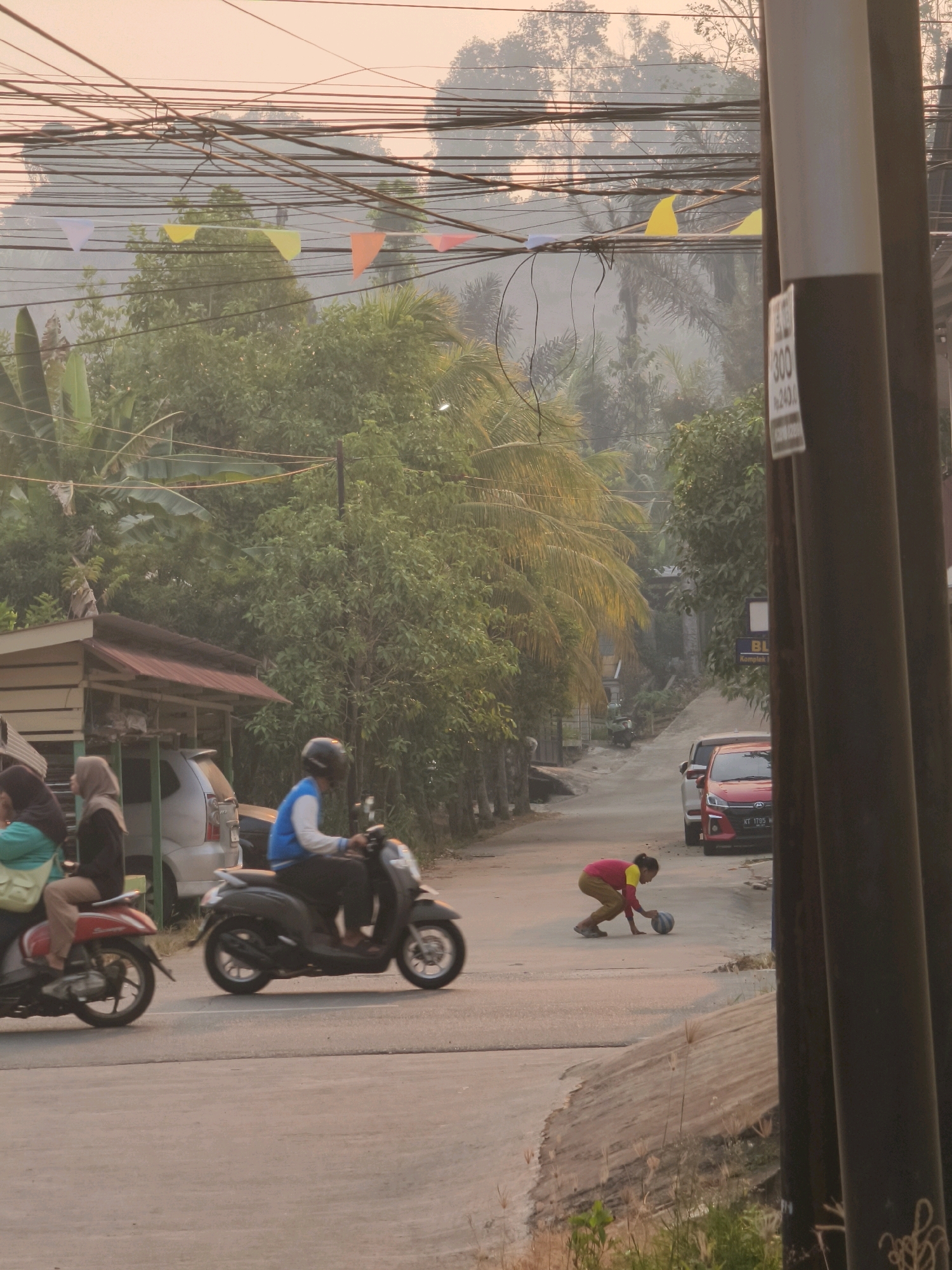 In Samarinda, Indonesia, a street scene unfolds with a gentle, hazy light characteristic of dusk or dawn. The road, paved with asphalt, curves gently uphill, flanked by lush green foliage and palm trees. Numerous electrical wires crisscross overhead, adorned with small, colorful pennant flags.
On the road, a person wearing a blue jacket and a black helmet rides a dark-colored scooter. Behind them, a red car is parked. To the left, another scooter carries two passengers, one in a light green shirt and the other in a dark top and hijab, both partially obscured by the scooter.
In the middle of the road, a child dressed in a pink and yellow outfit is crouched down, appearing to interact with a dark-colored ball. The child’s posture suggests they are about to pick it up or are in the process of playing with it.
A wooden structure with a red roof, possibly a small shop or shelter, is visible on the left side of the street. Utility poles stand on the right, one with a sign displaying the numbers "300" and "243.0". The overall atmosphere is tranquil, with a slight haze softening the edges of the landscape.