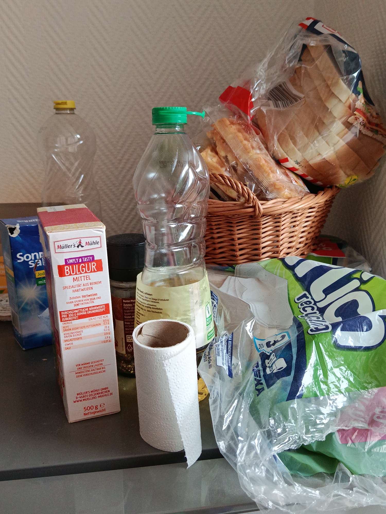 The image depicts a close-up view of various household items arranged on a dark gray surface, possibly a countertop. In the foreground, a roll of white paper towel is partially visible on the right. To the left of the paper towel, a cardboard box with red and white labeling, identified as "Müller's Mühle BULGUR MITTEL," is prominent. Adjacent to this box, to its left, is a blue cardboard box with white lettering, partially obscured, labeled "Sonn Salz," and appears to be a salt container.
Behind these items, a clear plastic bottle with a green cap is visible, containing a light-colored liquid. To its left is another clear plastic bottle with a yellow cap. In the background, a woven basket holds a plastic-wrapped package containing sliced bread. A large, crinkled green and blue plastic bag is on the right side of the frame, its contents unclear.
The overall environment is dimly lit, with a textured, neutral-colored wall or surface serving as the backdrop. No people are present in the image, and no actions or interactions are occurring. The arrangement suggests items that might be found in a kitchen or pantry. The city of Halbe, Germany is not depicted or referenced in this image.
