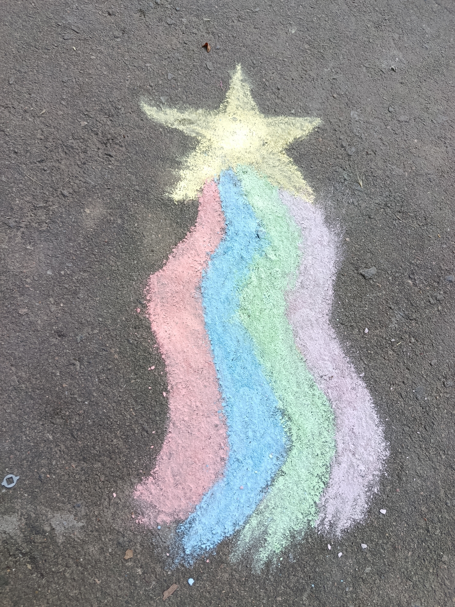 A chalk drawing of a shooting star with a rainbow trail is depicted on asphalt. The shooting star is yellow, and the rainbow trail consists of pink, light blue, green, and lavender stripes. The drawing appears to be created on a sunny day, as indicated by the bright colors and the lighting on the asphalt. The texture of the asphalt is rough and dark gray, with some small stones and debris visible. The image is taken from a slightly elevated angle, looking down at the drawing. There is no visible text in the image.