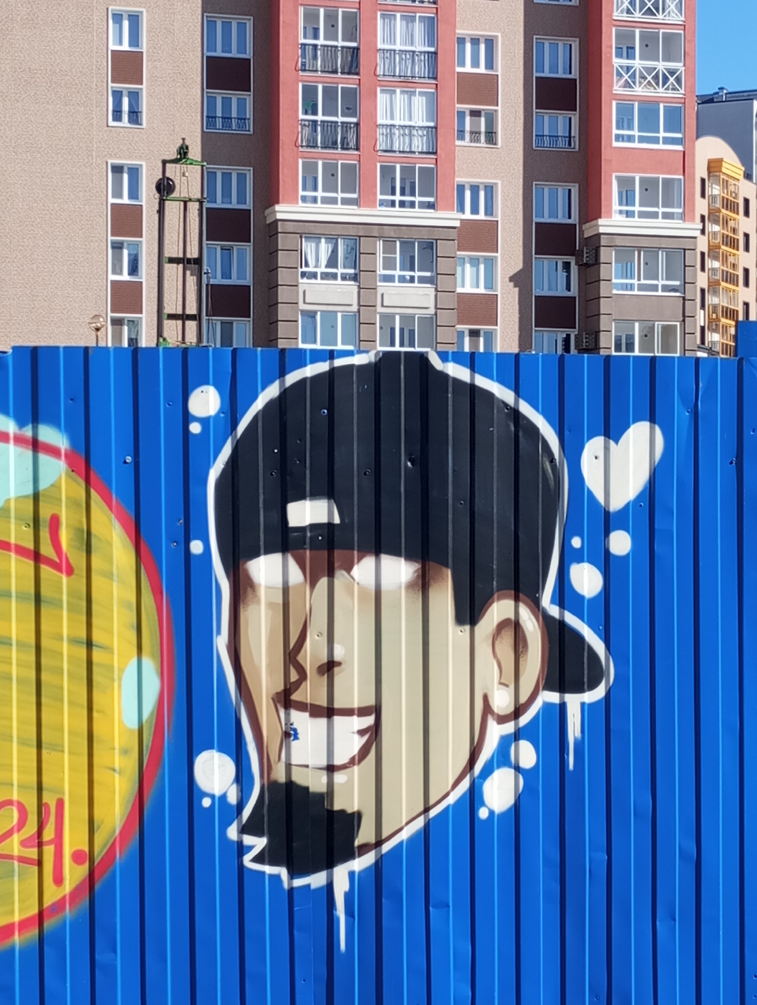 An outdoor, eye-level shot captures a vibrant graffiti artwork on a blue corrugated metal construction fence, with modern residential buildings forming the background under clear daylight.
The dominant feature in the foreground is a large, stylized cartoon head painted on the blue fence. The head depicts a person with light skin, wearing a black backward baseball cap. The face has strikingly white, pupil-less eyes, a wide, open-mouthed smile revealing white teeth, and a small, pointed goatee. White circular bubbles and a white heart shape float around the head, suggesting thoughts or emotions. To the left of the head, partially visible on the fence, is a segment of a yellow circle outlined in red, with red numbers "24." painted on it. The blue fence itself is corrugated, showing vertical ridges and shadows, indicating its texture and the bright sun.
In the mid-ground and background, several multi-story buildings are visible. The central and left buildings have a mix of beige and reddish-brown facades, interspersed with numerous windows, mostly white-framed. Some sections feature balconies with black or white railings, including some with decorative white cross-patterns. A tall, slender green metal structure, possibly part of a construction lift or scaffolding, stands against the beige building on the left. To the far right, another building with a distinctive yellow-brown exterior and many smaller windows can be seen. The bright sunlight casts distinct shadows, enhancing the depth and details of the scene. The overall impression is that of an urban or suburban development area, possibly under construction, brightened by street art.