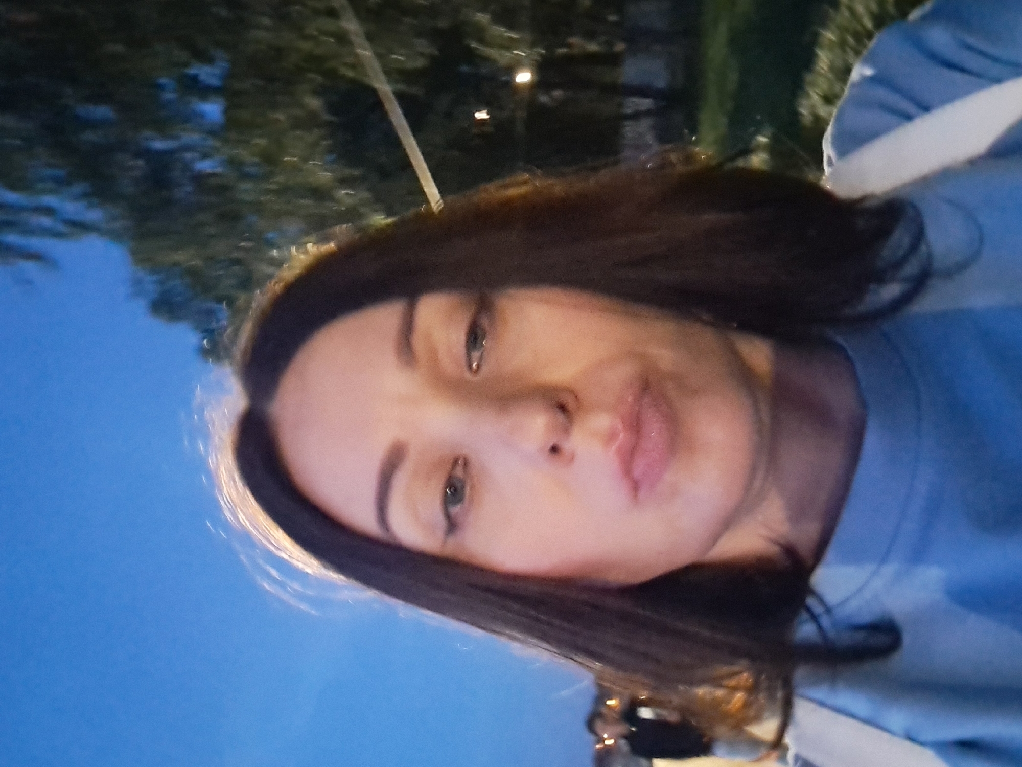 A selfie taken at dusk in Irkutsk, Russia, shows a woman with dark hair and light skin. She is wearing a light blue sweatshirt and a white strap across her shoulder. Her lips are pursed, and she is looking directly at the camera. In the background, trees and streetlights are visible, suggesting an outdoor urban setting. A blurry figure of a person is visible to the left of the frame. The sky is a deep blue, indicating twilight.