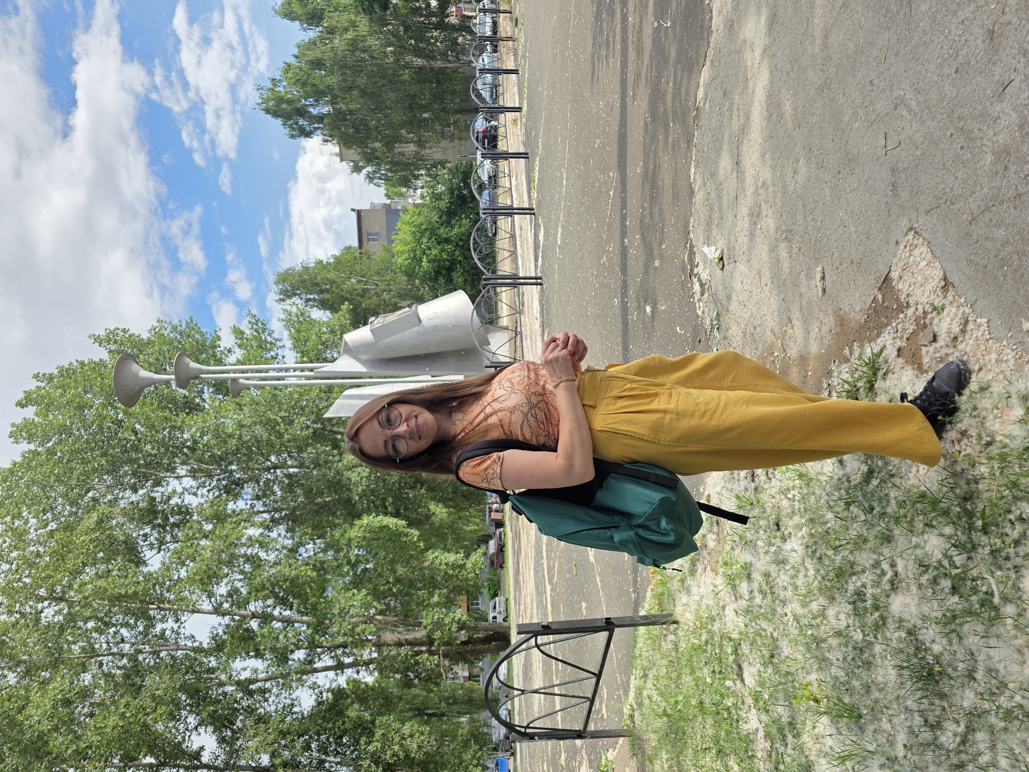 A full-body, eye-level shot shows a woman standing on a paved area outside, with a patch of grass to her left covered in white fluff. She is facing the camera, smiling slightly, and looking to her right. The woman has long, brown hair, is wearing glasses, a short-sleeved patterned top, and yellow wide-leg pants. A dark green backpack is slung over her right shoulder. To her left is a black metal fence with a decorative arched top. In the background, there is a modern metal sculpture on a pole with three large, horn-like structures. Behind the sculpture are several tall, leafy trees. Further in the background, beyond a row of similar black fences, are parked cars and apartment buildings. The sky above is blue with scattered white clouds. The ground in the foreground is a mixture of pavement and grass with white fluff. The overall scene appears to be a public space in the city of Chaikovsky, Russia, likely during the summer.