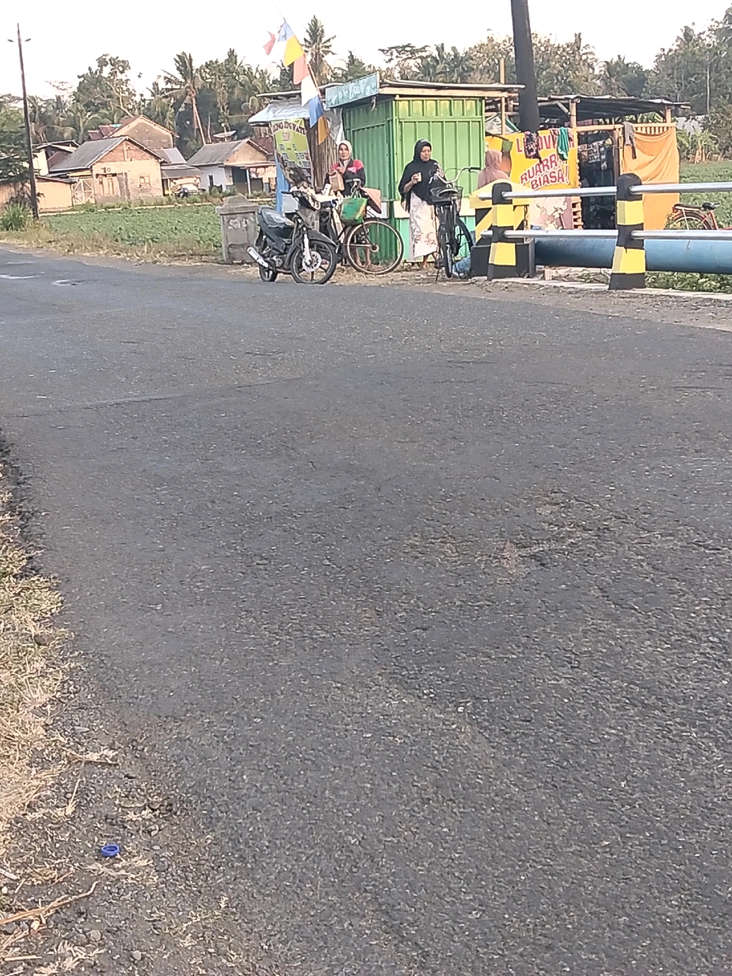 A street-level, eye-level outdoor photograph captured in Megulung Kidul, Indonesia. The image shows a paved road in the foreground with dry grass and some debris on the left side, including a blue bottle cap. In the middle ground, there is a small roadside stall with a green metal structure and a yellow banner displaying text that reads "LUVV RUARR BIASA." Two women, one wearing a dark hijab and the other a lighter-colored headscarf, are standing near the stall. One woman is holding a black bag, and the other appears to be holding a cup. Two bicycles are parked near them, one in front of the woman in the dark hijab and another to the right. A black motorcycle is parked to the left of the bicycles. In the background, there are several houses with red-tiled roofs and palm trees, suggesting a rural or semi-urban environment. A blue pipe runs horizontally across the mid-background, supported by yellow and black striped pillars. The sky is overcast.