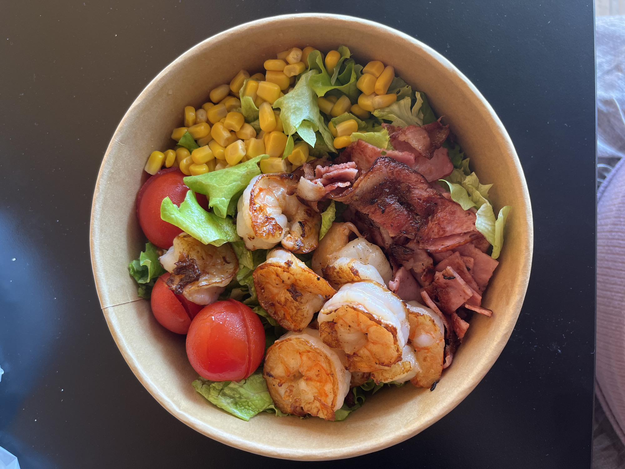 A high-angle, close-up shot shows a salad in a tan paper bowl. The salad is composed of lettuce, corn kernels, cherry tomatoes, cooked shrimp, and sliced bacon. The bowl rests on a dark surface next to what appears to be a lilac blanket or fabric. There's no indication of the location being Varna, Bulgaria.