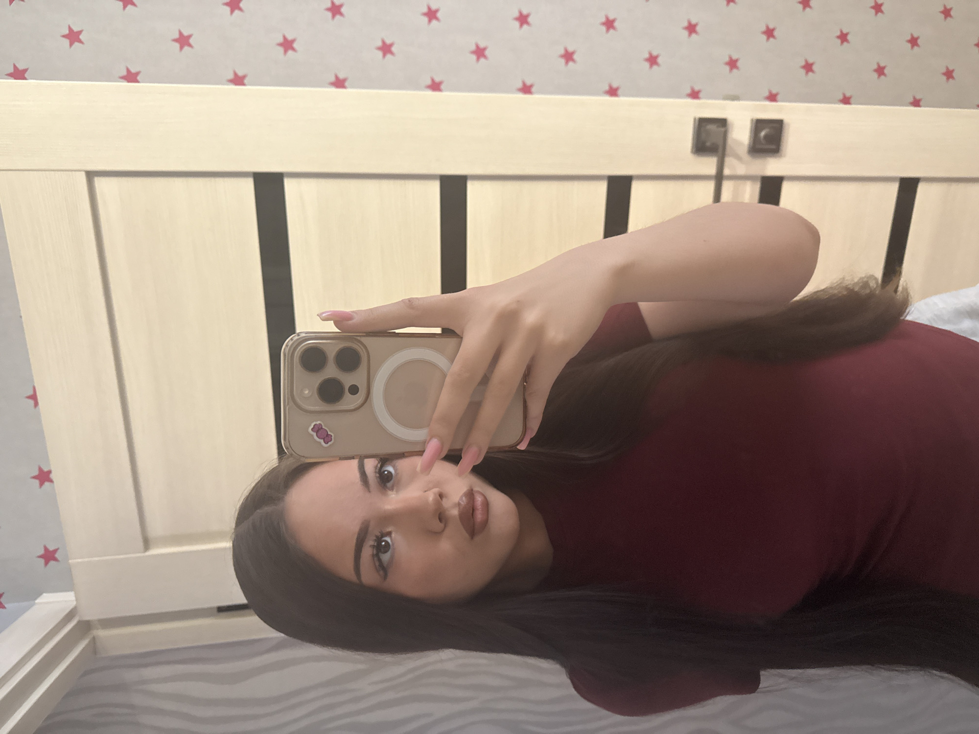 The image captures a rotated mirror selfie of a young woman, likely taken in a bedroom in Naberezhnye Chelny, Russia. The woman, with long dark hair, is lying on her side, wearing a dark red or maroon top. She holds a smartphone with a light-colored case, featuring a prominent circular design and a small cartoon candy sticker, covering part of her face. Her eyes are wide and focused on the camera, and her lips are slightly pursed in a playful or inquisitive expression. Her long nails are painted a light pink. The background shows a light wooden headboard with darker vertical accents. Above the headboard, a light-colored wallpaper patterned with numerous red stars is visible. Two dark, square-shaped fixtures, possibly lights or electrical outlets, are mounted on the headboard. A striped bedsheet or blanket is visible beneath her.