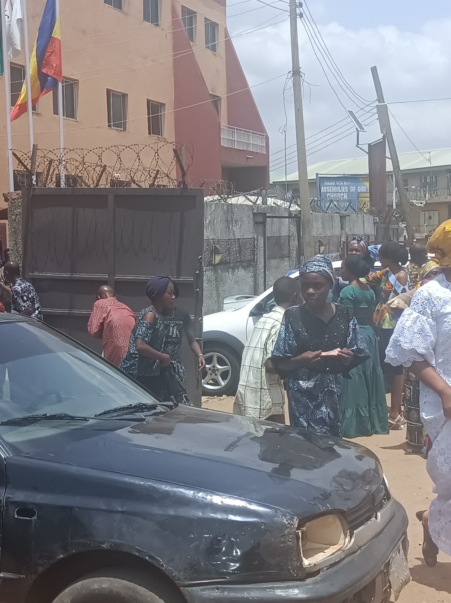 The image depicts a street scene in Dadi Riba, Nigeria. In the foreground, a black car with front-end damage, specifically a missing headlight and bumper damage, is partially visible. Behind the car, a group of people, predominantly women, are gathered. They are dressed in various traditional and contemporary clothing, including colorful fabrics, head wraps, and lace garments. Behind this group and on the left is a closed metal gate topped with barbed wire. Beyond the gate stands a peach-colored building with flags of different nations flying. In the background, across the street, a sign for the Assemblies of God Church is visible amidst other buildings and utility poles. The overall environment suggests a busy street scene with a mix of pedestrian and vehicular traffic, near a church and in front of a gated compound.