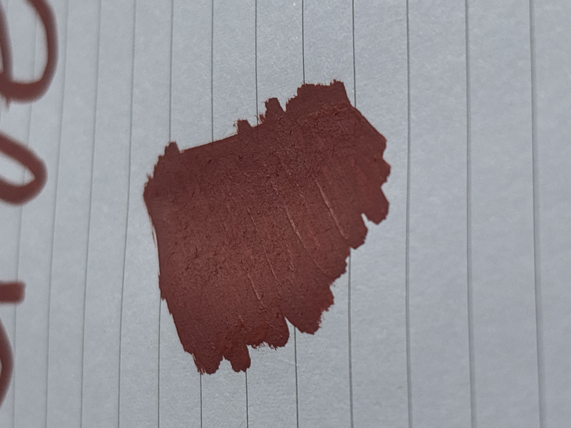 A close-up, slightly angled shot of a swatch of reddish-brown lipstick is shown on a piece of lined paper. The lipstick swatch is textured and appears matte, with visible brushstrokes. The paper has horizontal blue lines. Above the lipstick swatch, parts of red lettering are visible, possibly part of the word "love." There are no people, other objects, or indications of a specific location in Saint Petersburg, Russia.