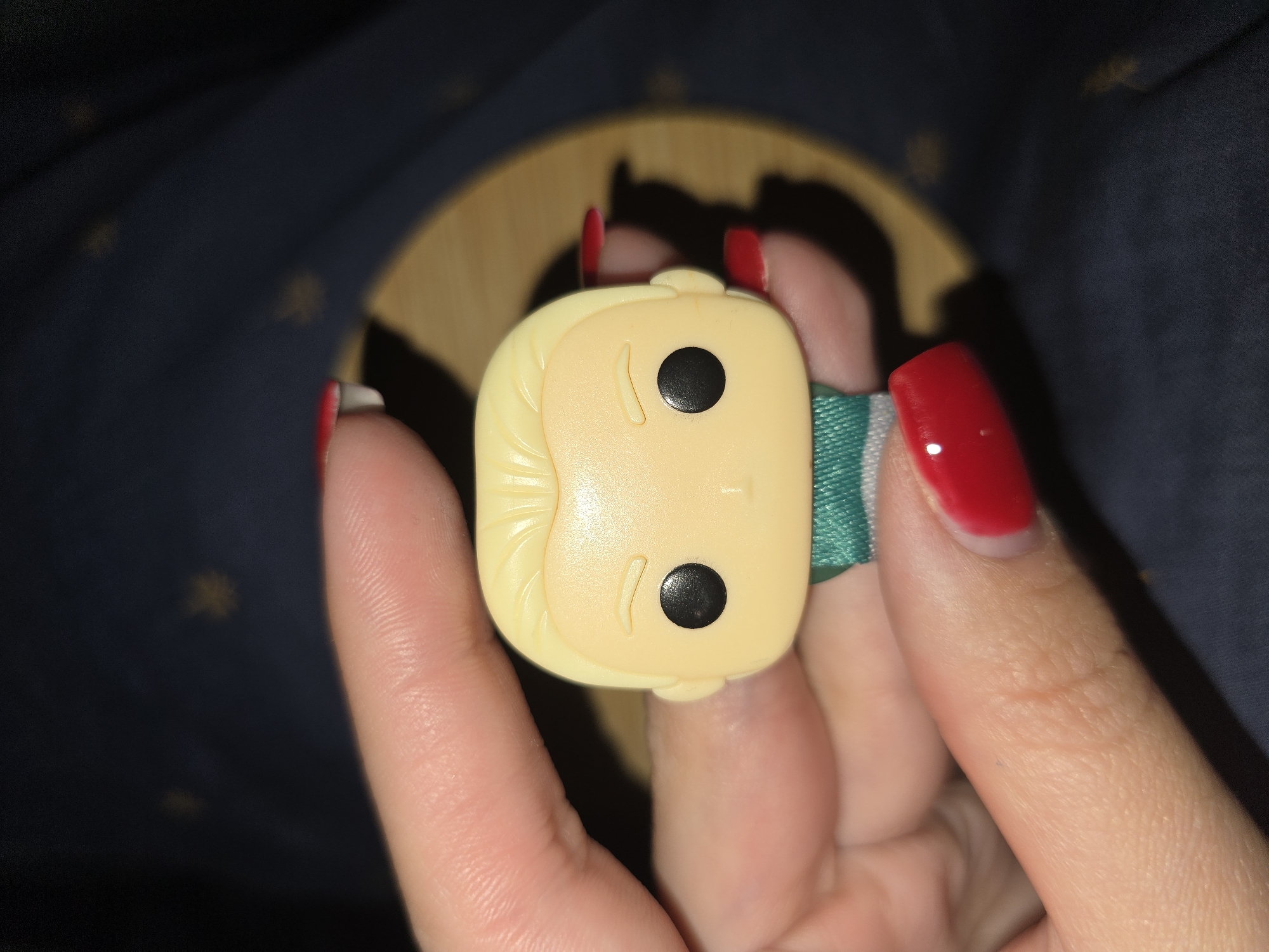 The image is a close-up, overhead shot of a person's hand holding a Funko Pop accessory. The accessory is a small, stylized head of a character, made of light yellow plastic. It has two black dots for eyes and a molded line indicating a nose. The character has blonde hair with sculpted strands. A thin, green and white striped band wraps around the accessory.
The hand holding the accessory has red-painted fingernails. The skin texture of the fingers is visible, with lines and creases. The background is dark, with some blurry yellow star-like shapes and a circular wooden object out of focus. The lighting suggests an indoor setting, possibly at night or in low light conditions. No visible text is present in the image. The overall impression is of a casual, personal snapshot.