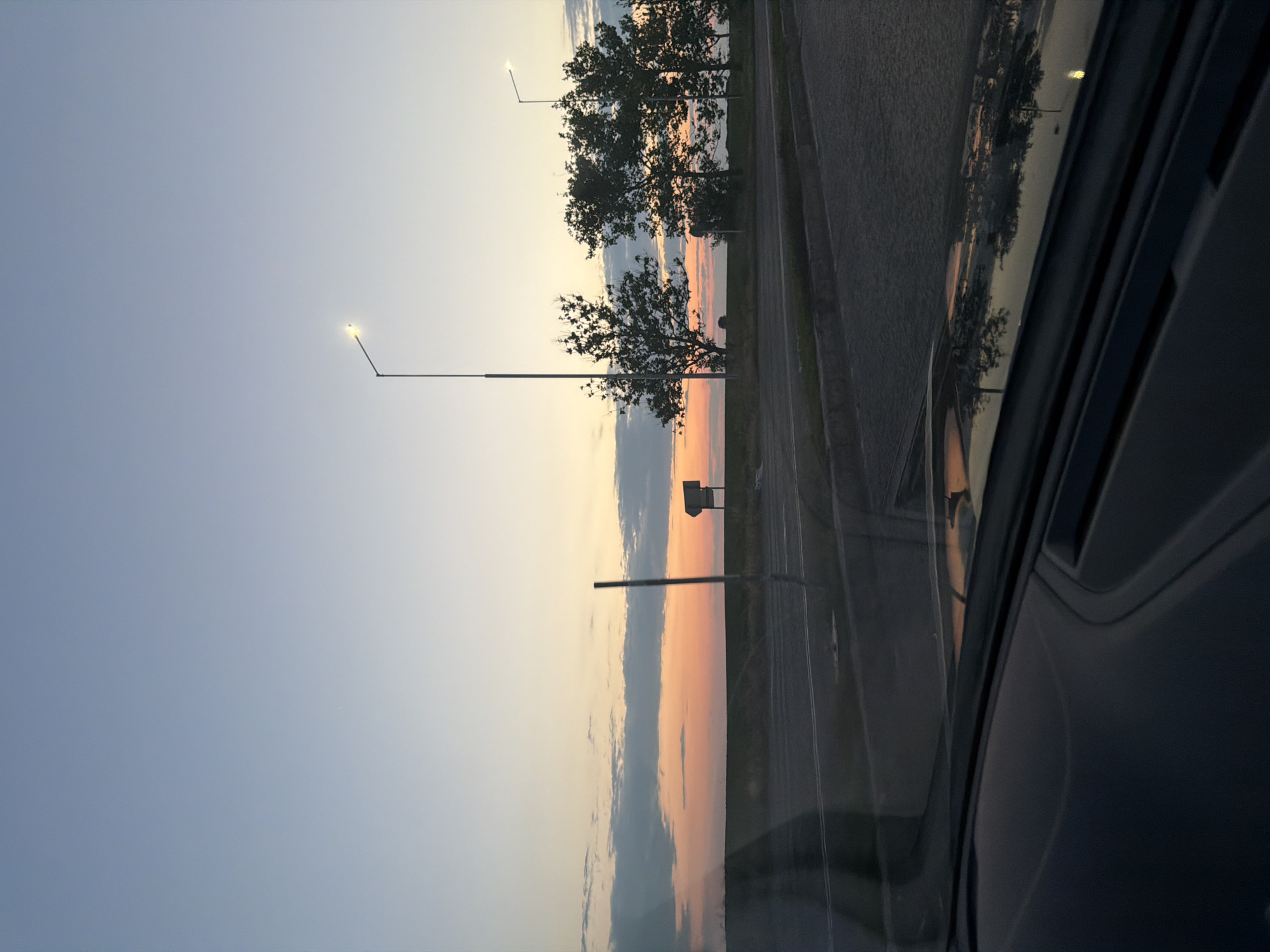 The image presents an outdoor scene at twilight, viewed from the interior of a vehicle looking forward through the windshield, set in the vicinity of Dolno Ablanovo, Bulgaria. The sky displays a gradient of colors, transitioning from a pale blue at the zenith to yellow, orange, and finally a vibrant pink-orange hue at the horizon, characteristic of either sunrise or sunset. Scattered clouds, some with darker undersides, are visible within the warmer segments of the sky. A small, bright white celestial object, likely a star or planet, is discernible in the upper left quadrant of the sky.
Two streetlights are illuminated within the scene. A prominent streetlight with a bright white lamp is positioned to the right of the image's vertical center. A second, smaller illuminated streetlight is visible further to the right, partially obscured by tree foliage.
The midground features a flat, dark landscape, likely agricultural fields, extending to the horizon. Several deciduous trees, appearing sparsely foliated, are silhouetted against the colorful sky. A dark vertical pole stands in the left-midground, and a small, indistinct, light-colored sign or structure is visible on the horizon to its right.
The immediate foreground shows a paved road surface, dark grey with lighter markings, and a distinct curb separating it from a darker shoulder area. The top portion of a dark-colored vehicle dashboard occupies the bottom of the frame. The windshield exhibits significant reflections of the external environment, including the colored sky, trees, and streetlights, particularly noticeable in the lower right section of the image. No people or animals are visible.