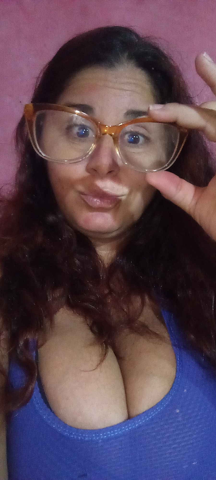 A woman with long, auburn-colored hair is wearing amber-colored glasses and a blue tank top. She is holding the glasses up to her face with her right hand. The background is a mottled pink.