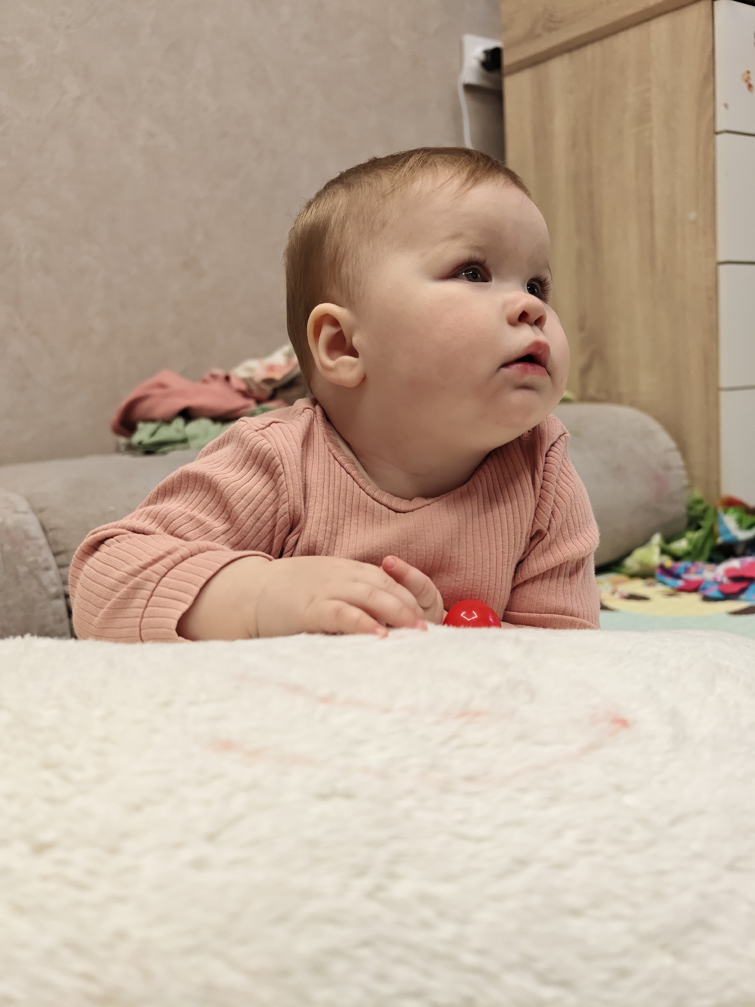 The image depicts an infant engaged in tummy time indoors. The infant, with light brown hair and fair skin, is wearing a long-sleeved, pink ribbed top. The child is positioned on a white, soft, textured surface, possibly a rug or blanket, looking upwards and to the right with a slightly open mouth. A small, red, dome-shaped object is visible beneath the infant's right hand.
In the background, to the left, is a grey upholstered sofa or bed with a pile of folded clothes in shades of pink, green, and multi-colored patterns. To the right, a light wood-grained cabinet or dresser with white drawers is partially visible. An electrical outlet with a white plug is mounted on a light-colored wall between the sofa and the cabinet. The general environment suggests a residential indoor setting. The specific location within the city of Chaikovsky, Russia, is not discernible from the visual information provided in the image.