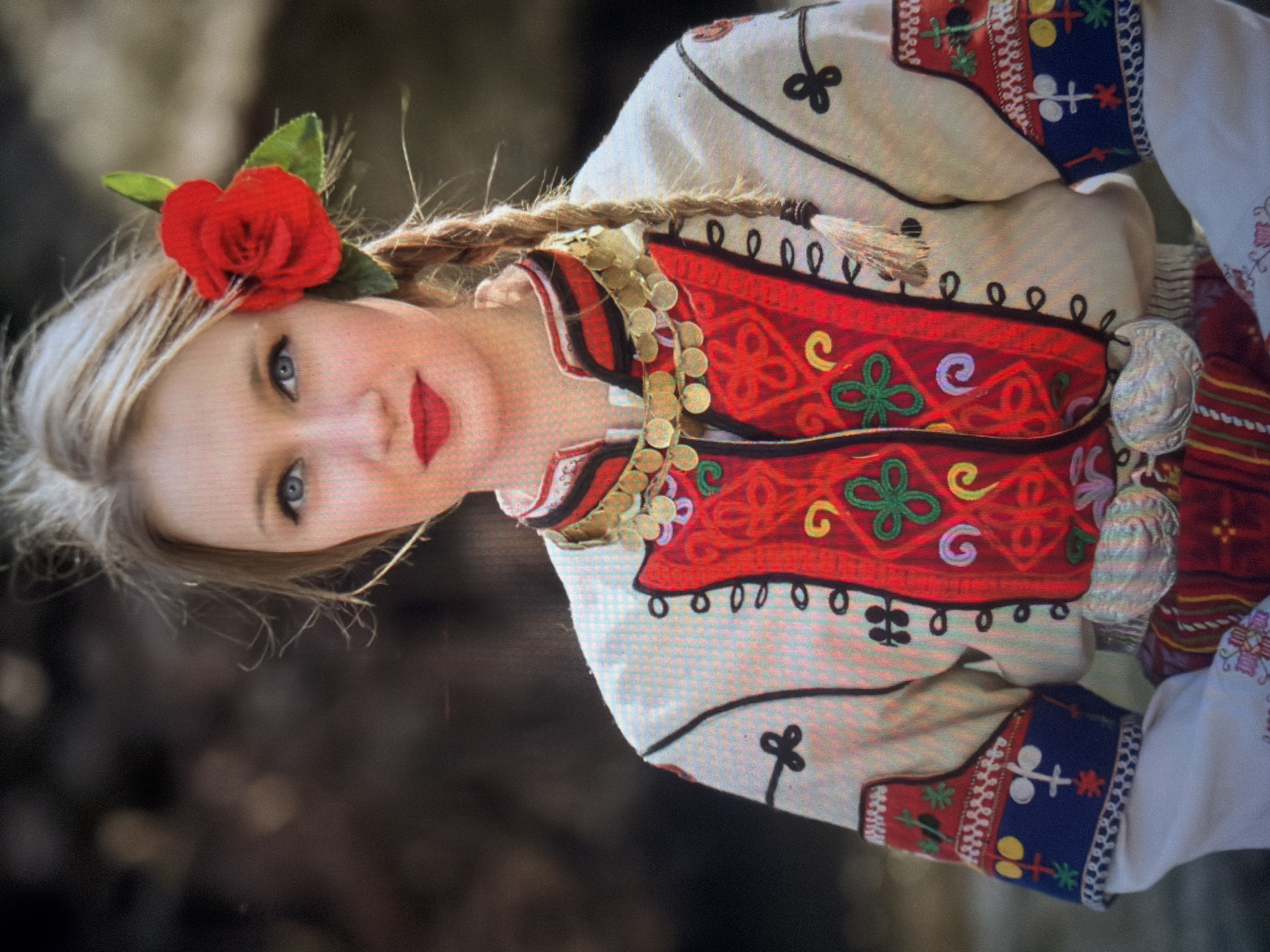 A young woman is depicted in a close-up, head-on shot in Shumen, Bulgaria, showcasing traditional Bulgarian folk attire. She has fair skin, blue eyes, and light blonde hair styled with a single braid extending over her right shoulder. A bright red artificial rose with green leaves is tucked behind her right ear. Her lips are painted with red lipstick. She wears a white long-sleeved shirt featuring intricate embroidery on the cuffs, characterized by a blue band with red, green, yellow, and white geometric and symbolic motifs. Over the shirt, she wears a red vest heavily embroidered with yellow, green, and purple swirling patterns and geometric designs, outlined with black trim and small black loops down the front opening. A multi-strand necklace of gold-colored coin-like medallions encircles her neck. A wide, patterned belt, possibly red and white striped, is visible around her waist, secured by large, ornate silver-colored buckles or plaques at the front. The background is softly blurred with dark, earthy tones, suggesting an outdoor setting.