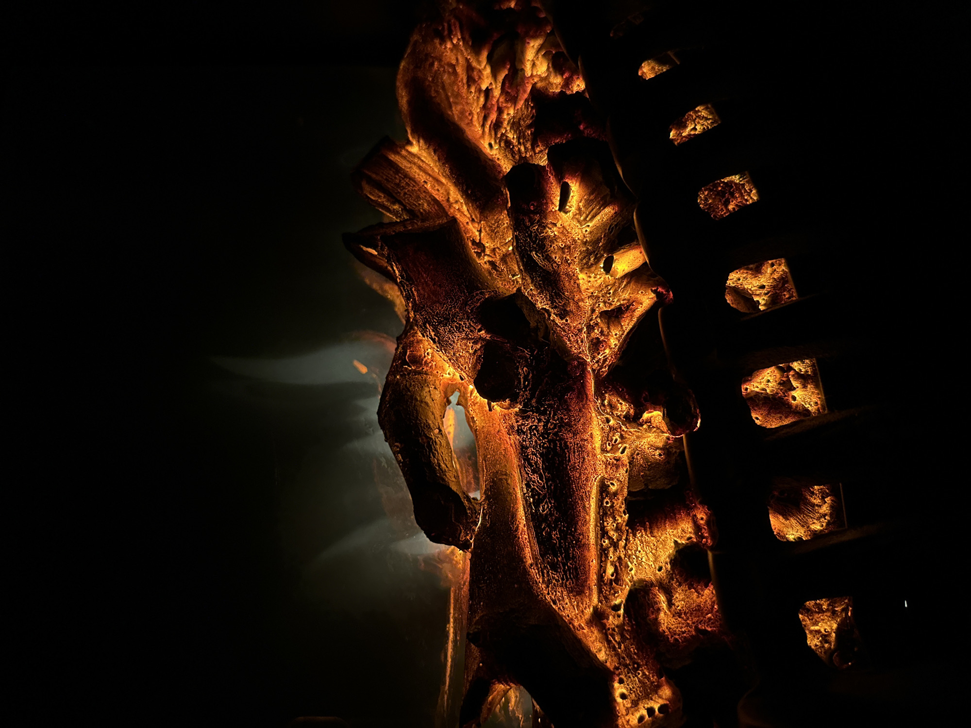 The image depicts a close-up view of artificial logs and glowing embers inside a dark fireplace. The logs are textured and appear to be made of a material that simulates burnt wood. They are illuminated from within by a warm, orange-yellow light, creating a realistic effect of embers. The background is completely black, emphasizing the glow of the artificial fire. The scene is captured in a way that highlights the dramatic contrast between light and shadow, giving it an intimate and cozy feel. There are no people or animals present in the image. The overall impression is one of warmth and simulated fire.