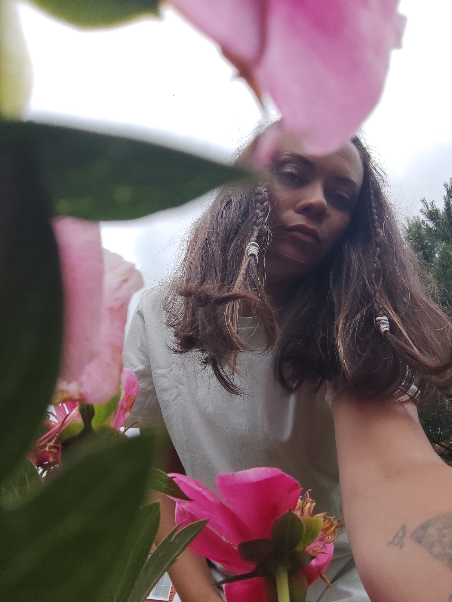 A low-angle, close-up photograph taken in the city of Фоки, Russia, captures a person framed and partially obscured by out-of-focus vibrant pink flowers and green foliage in the immediate foreground. The individual, appearing female, has long, dark brown hair styled with several braids accented by light-colored wraps or beads. She wears a light-colored top and looks directly at the camera with a neutral expression. A partial tattoo is visible on her left forearm. The background features an overcast, light grey sky and dark green coniferous trees to the right. A portion of a reddish-brown building with a white-framed window is discernible through the leaves on the lower-left side.