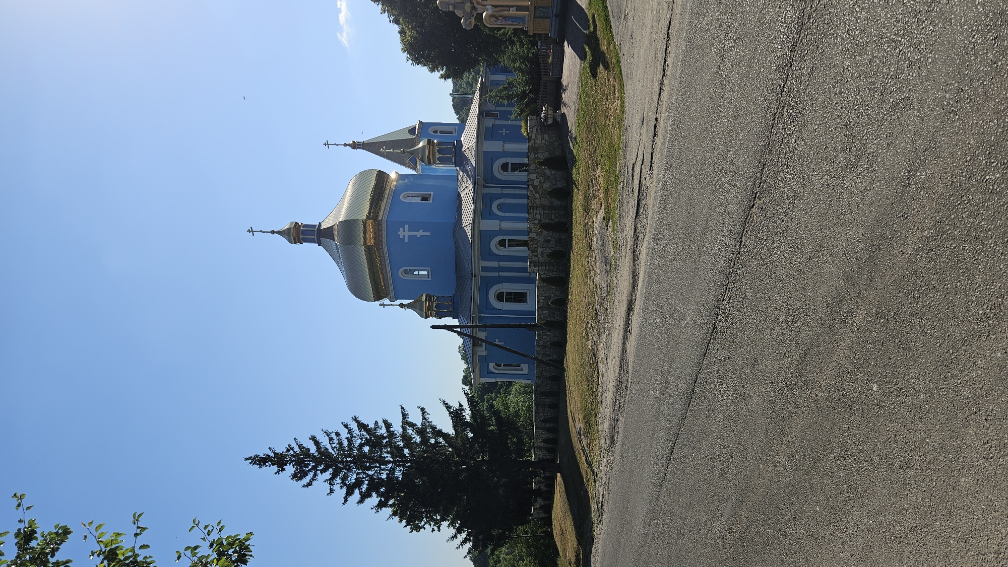 The image depicts a blue Eastern Orthodox church with golden domes. The church has a prominent central dome and a smaller dome on the left, both topped with crosses. The building features arched windows and decorative elements. It is situated in an outdoor setting, with a paved road in the foreground and a grassy area with some trees and foliage to the side and behind the church. A tall evergreen tree stands in front of the church. The sky is clear and blue, suggesting a sunny day. The orientation of the image is rotated 90 degrees clockwise. There is no visible text in the image. The scene appears to be in a rural or semi-rural area. The time of day is likely daytime, given the bright lighting and clear sky.