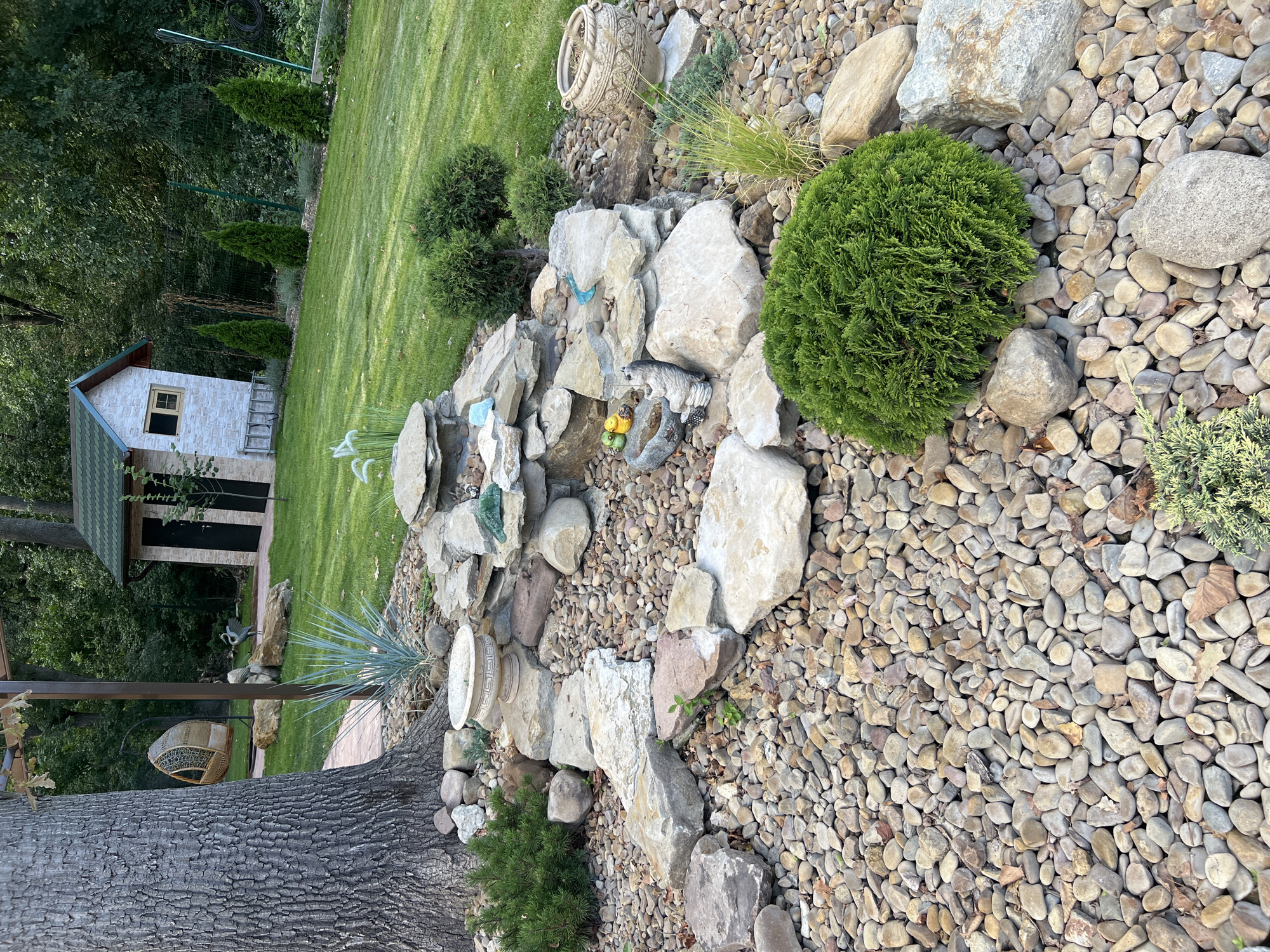This is an outdoor scene of a backyard garden in Kitskany, Moldova. The foreground is dominated by a rock garden featuring a variety of stones and pebbles, with several small green bushes strategically placed throughout. A large tree trunk is visible on the left side of the image, with a hanging chair suspended from a metal frame. In the midground, a multi-tiered rock formation appears to be a small waterfall or water feature, with a light blue accent visible on one of the rocks. Two small yellow duck figurines and a gray ceramic figurine of a cat are placed on the rocks. Behind the rock garden, a well-maintained green lawn stretches towards the background. A small, white house with a green roof, resembling a garden shed or playhouse, is situated on the right side of the lawn. A metal ladder leans against the side of the house. Several slender, green evergreen trees are planted in a row behind the house. The background consists of dense green trees and foliage, indicating a wooded area beyond the garden. The overall impression is of a cultivated and decorative outdoor space.