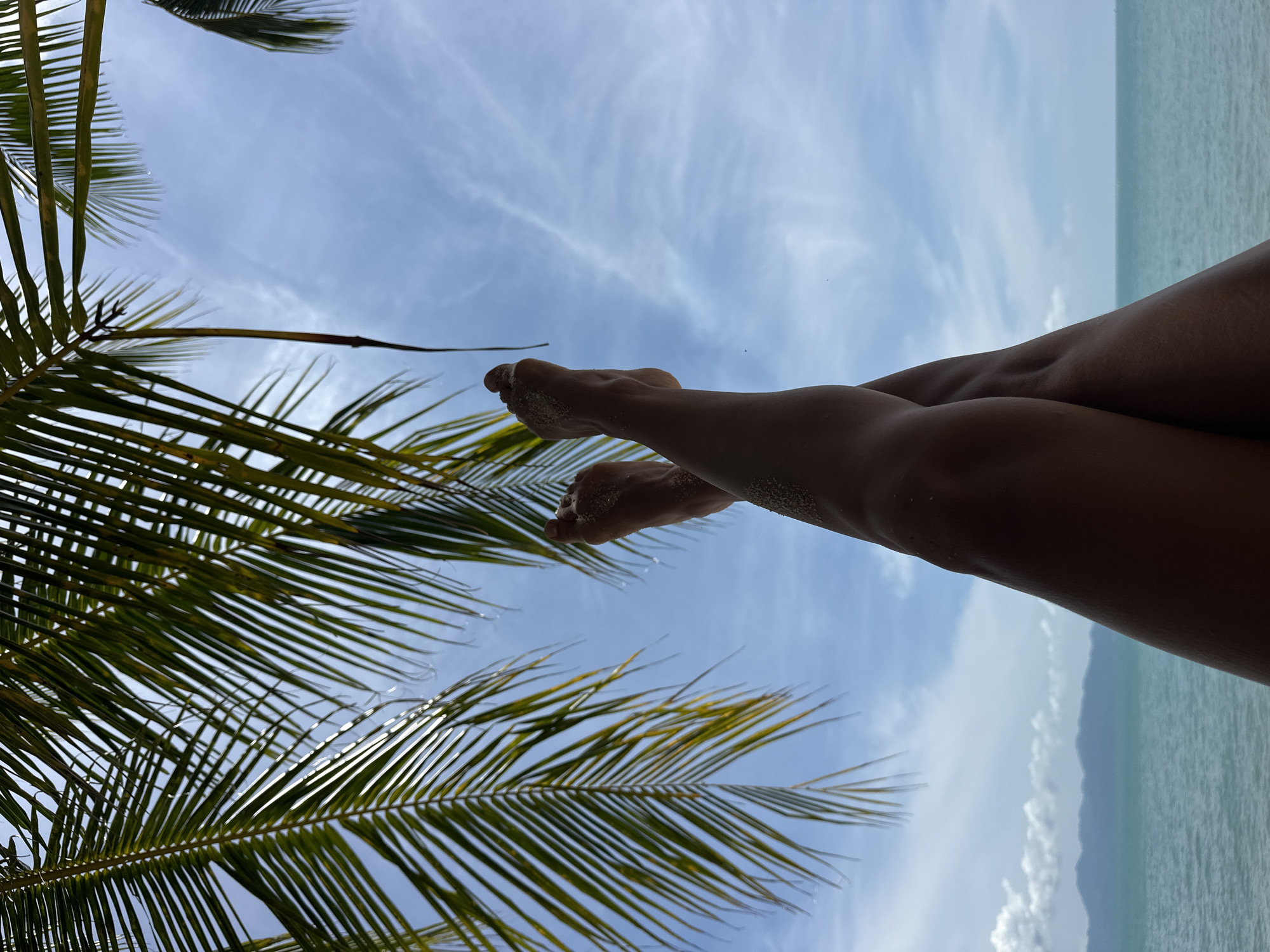 The image, taken from a low angle perspective, shows a pair of legs and feet crossed against a backdrop of a blue sky with scattered clouds. Palm fronds are visible in the upper portion of the frame, casting shadows. The ocean and a distant island or landmass are visible at the bottom, indicating a tropical coastal setting. The legs appear tanned and are dusted with sand, suggesting the person is at a beach. The overall mood is one of relaxation and leisure. The specific location in Ko Samui District, Thailand, is not discernible from the image itself, but the scenery is consistent with the island's known tropical environment.