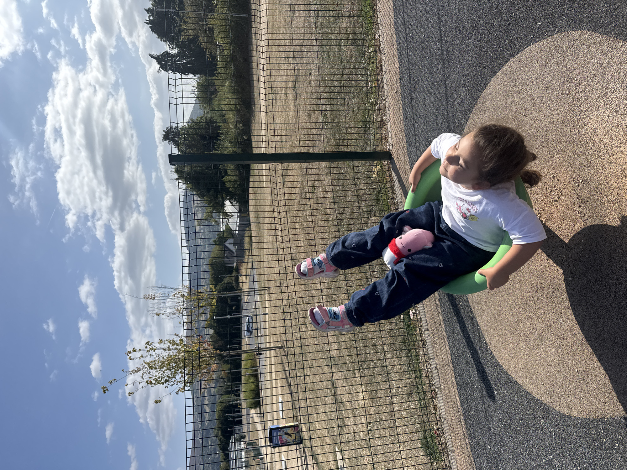 A young girl with blonde hair is lying on her back inside a green, circular spinning disc in a playground. She is wearing a white long-sleeved shirt with a graphic of a cartoon character, dark blue jeans, and pink sandals. A small pink plush toy with a red hat is tucked between her legs. The playground has a speckled grey and brown rubberized surface with a large circular area where the disc is situated. A black chain-link fence is visible behind the girl. Beyond the fence, dry grass, trees, and buildings are present, with a hilly landscape and a blue sky with scattered clouds in the background. The scene appears to be a sunny day. The location is the playground adjacent to the Clonmel Library and Town Hall, in Clonmel, County Tipperary, Ireland.