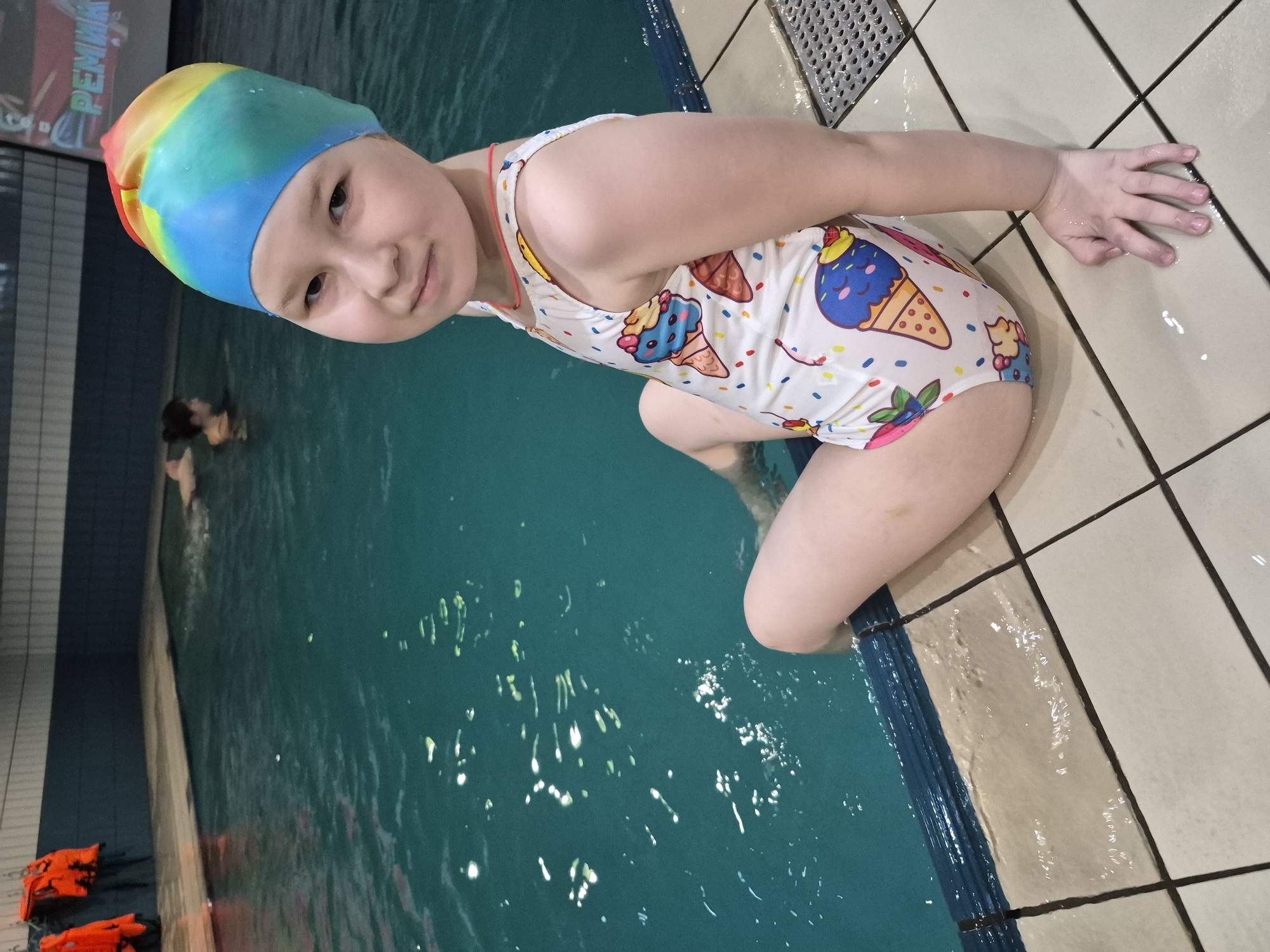 A young girl wearing a rainbow-colored swim cap and an ice cream-patterned swimsuit is sitting at the edge of a swimming pool. She has one hand resting on the tiled poolside and is looking towards the camera. The pool water is turquoise. In the background, there is a person in the water and orange life jackets are visible on the pool's edge. The location is possibly a swimming pool in Tobolsk, Russia, based on image metadata.