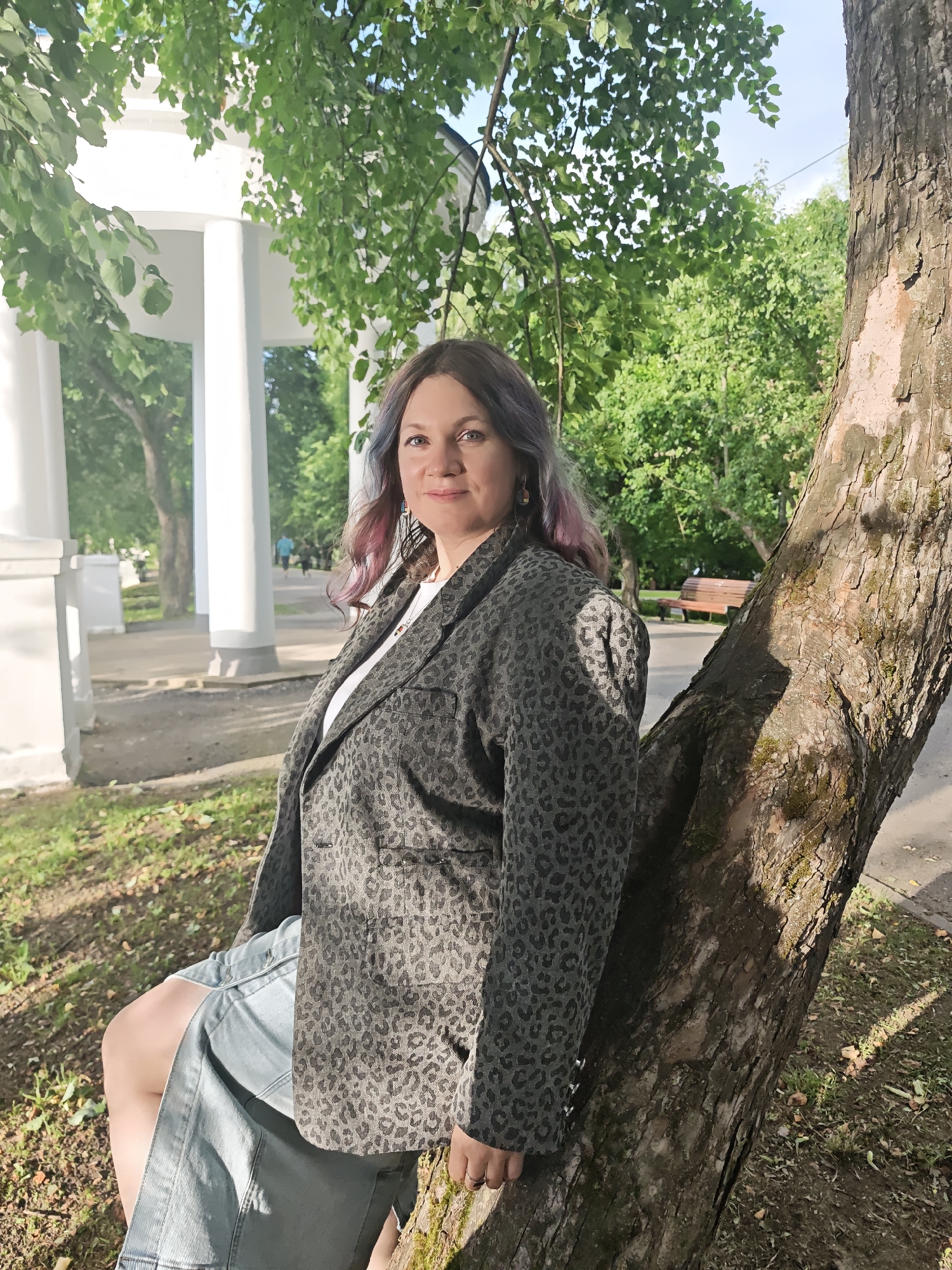 A woman with long, wavy hair streaked with purple and blonde poses in a park. She is wearing a grey leopard-print blazer over a white t-shirt and a light blue denim skirt. She is leaning against a large tree with rough bark, with her legs crossed. In the background, a white gazebo with columns is visible, surrounded by lush green trees. The sun is shining, casting dappled shadows on the ground, suggesting it is a pleasant day. The woman is looking at the camera with a slight smile, appearing relaxed and content. The scene is set in a park in Dubna, Russia, indicated by the prompt's location context. The time of day appears to be late afternoon due to the warm sunlight.