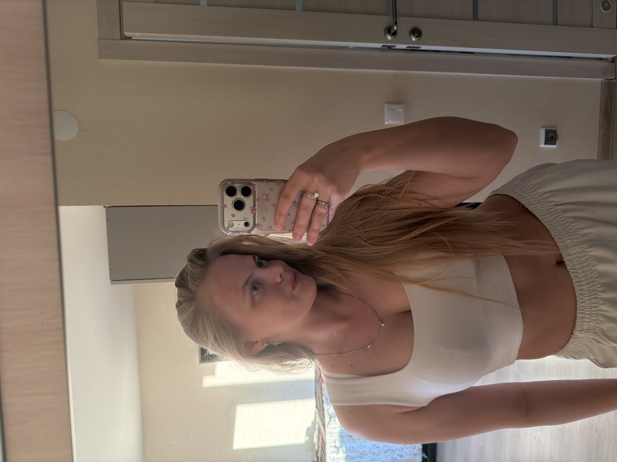 A young woman with long, straight, light blonde hair captures a mirror selfie indoors during the daytime. She is holding a smartphone in her left hand, which is encased in a clear cover adorned with small floral or cherry patterns. She wears two rings on her left ring finger, one of which features a small white heart design.
She is dressed in a plain white or light-colored sleeveless crop top that exposes her midriff, paired with light-colored, possibly cream or white, elastic-waist pants or shorts. A delicate silver-toned necklace with a small heart-shaped pendant is visible around her neck. Her gaze is directed towards the camera in her reflection, with a neutral to slightly pensive expression.
The background visible in the mirror shows an interior space with light-colored walls, possibly beige or off-white. Wooden door frames or trim are present, along with a circular fixture on the upper left wall and several electrical outlets or light switches. Natural light, likely from a window, illuminates the scene, suggesting it is daytime. The setting appears to be a modern residential space, such as a bedroom or hallway. No specific location cues are visible within the image itself, and no discernible text is present beyond the patterns on the phone case.