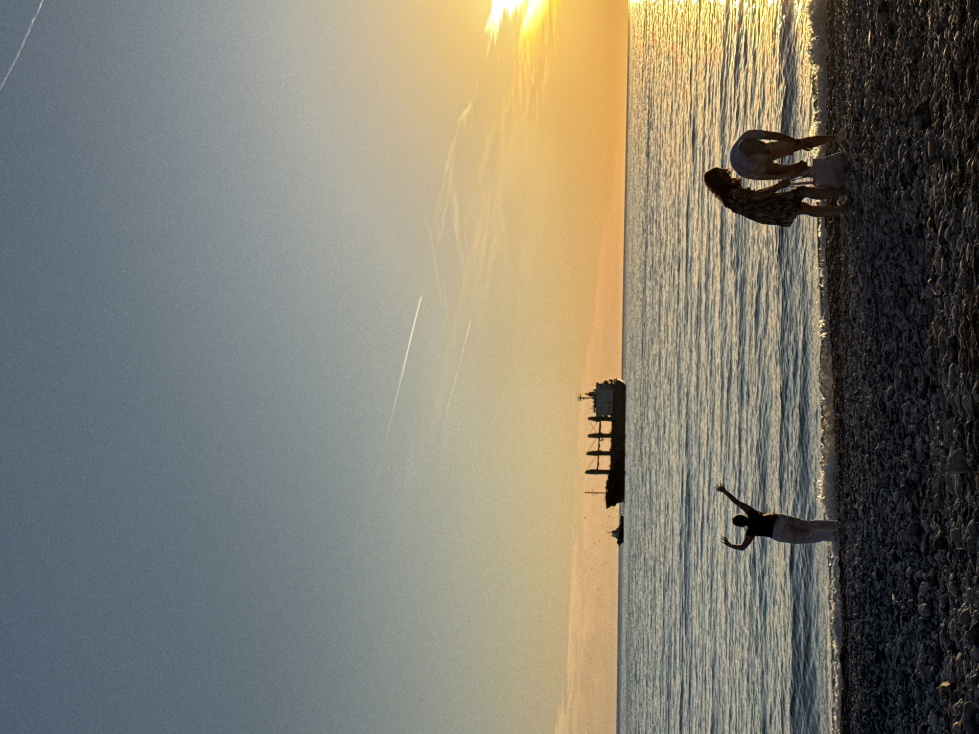 The image shows a beach scene at sunset in Bar, Montenegro. The sun is setting over the sea, casting a golden glow on the water and the sky. A large cargo ship is visible in the distance. Three figures are on the beach. A woman is standing near the water's edge with her arms raised. Two other figures are further down the beach bending over. The beach is made of pebbles.