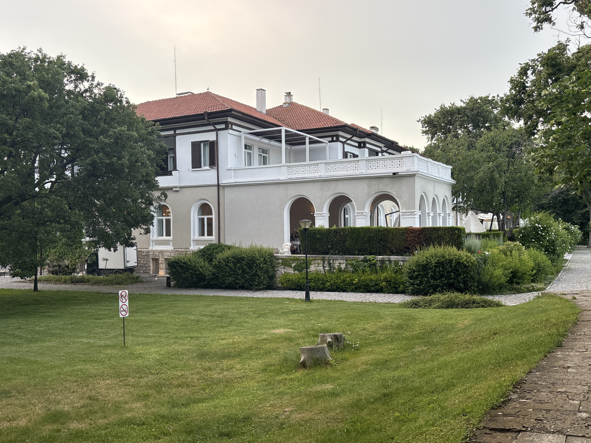 The image depicts a large, multi-story building with a red tile roof and white exterior walls, featuring arched windows on the ground floor and a white railing on the second-floor balcony. The building is surrounded by lush green trees and manicured bushes. A stone pathway runs along the right side of the image, adjacent to a well-maintained lawn. In the foreground, two tree stumps are visible on the grass, and a signpost with two circular prohibition symbols (one depicting a person, the other a cigarette) is planted in the lawn. A white truck is parked near the building to the left, partially obscured by a large tree. The sky is overcast, suggesting it is either early morning or late evening. The architectural style and the surrounding vegetation suggest a location in a park or a large estate within Varna, Bulgaria.