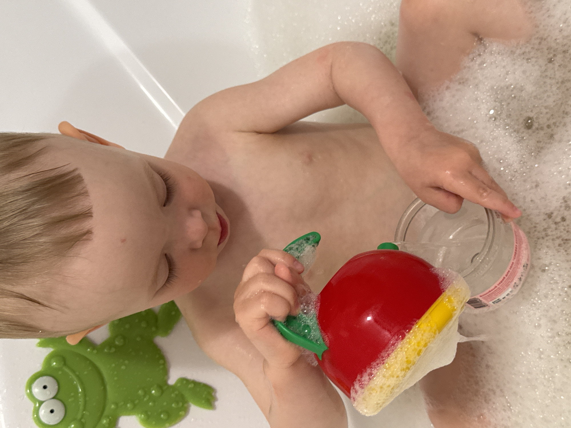 The media file shows a close-up, overhead view of a young child in a bathtub filled with foamy water. The child is fair-skinned with blond hair and is partially submerged in the bath. The child's face is mostly obscured, but their closed eyes and open mouth suggest they are relaxed and enjoying bath time.
The child is holding two bath toys. In their right hand, they grip a red, apple-shaped watering can with a yellow rim and green stem, which is also covered in foam. In their left hand, the child is holding a clear plastic container, possibly a bottle or a cup, from which water is being poured. The container has a pink label with some text visible, but it is difficult to read due to the water and foam. To the left of the child, a green rubber frog toy with large white eyes is partially visible.
The scene is brightly lit, with a strong light source creating reflections on the water's surface. The presence of soap bubbles indicates that the child is actively engaged in bathing. The overall mood appears to be one of childhood enjoyment and playful activity in a domestic setting, likely a bathroom. No specific time of day or weather cues are evident, but the location context provided is Chaikovsky, Russia.