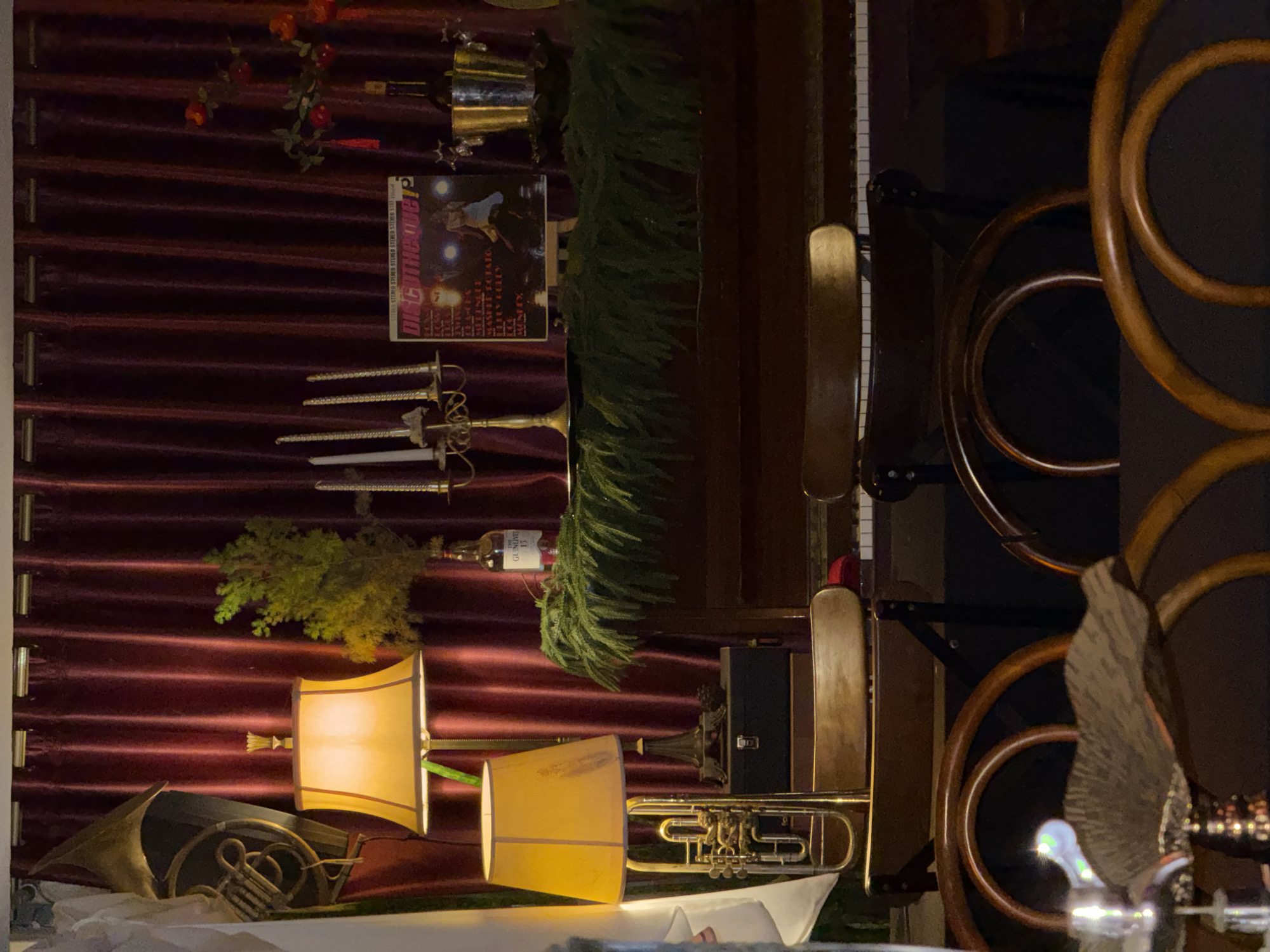 The image presents a scene within a dimly lit interior, likely a bar or a room designed with a vintage aesthetic in Lâm Viên, Đà Lạt, Vietnam. The backdrop is a red, pleated velvet curtain, against which several ornate objects are displayed. These objects include musical instruments such as a French horn and a trumpet, antique-style lamps with yellow shades, and decorative swords. A vinyl record and a bottle of Glenfiddich 15-year-old Scotch whisky are also noticeable. The entire arrangement has a theatrical quality. Additionally, there is a wooden structure with circular details. A decorative eagle can be seen in the lower right.