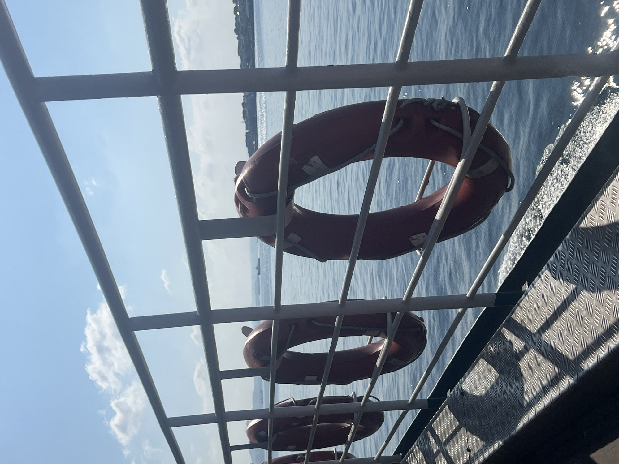 A medium shot captures a view from the deck of a ferry or boat navigating the waters of Prince Islands, Turkey. In the foreground, a metal railing divides the frame, behind which several orange life preservers are hung. The life preservers are circular with white markings and rope details. The deck itself is visible in the lower left, featuring a diamond-plate metal surface reflecting sunlight and casting shadows. The sea, a deep blue, occupies the middle ground, with gentle ripples and a white foam trail on the right side, indicating the boat's movement. In the distance, the shore of Prince Islands is visible, featuring a line of buildings and a church steeple against a bright, slightly hazy sky dotted with white clouds. A few distant ships are also discernible on the horizon. The lighting suggests it is a sunny day.