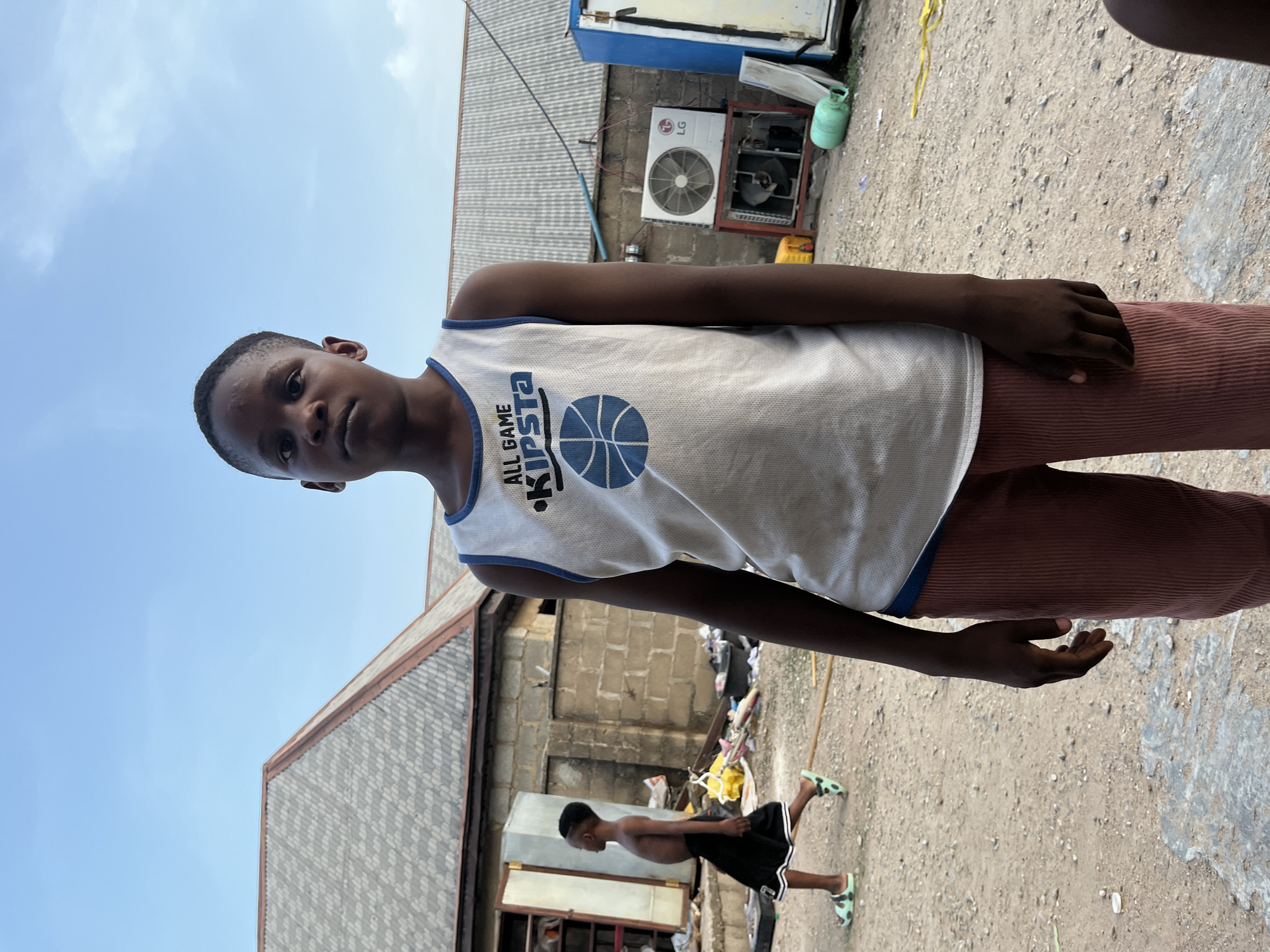 A young boy in a white tank top with the word "KIPSTA" and a basketball graphic stands facing the camera. He wears reddish-brown corduroy pants. Behind him, a second shirtless boy in shorts and sandals walks away. The background consists of buildings, including one with an LG air conditioner, and a dirt ground with debris. The scene appears to be located outdoors. The location of the image is Iddah, Nigeria.