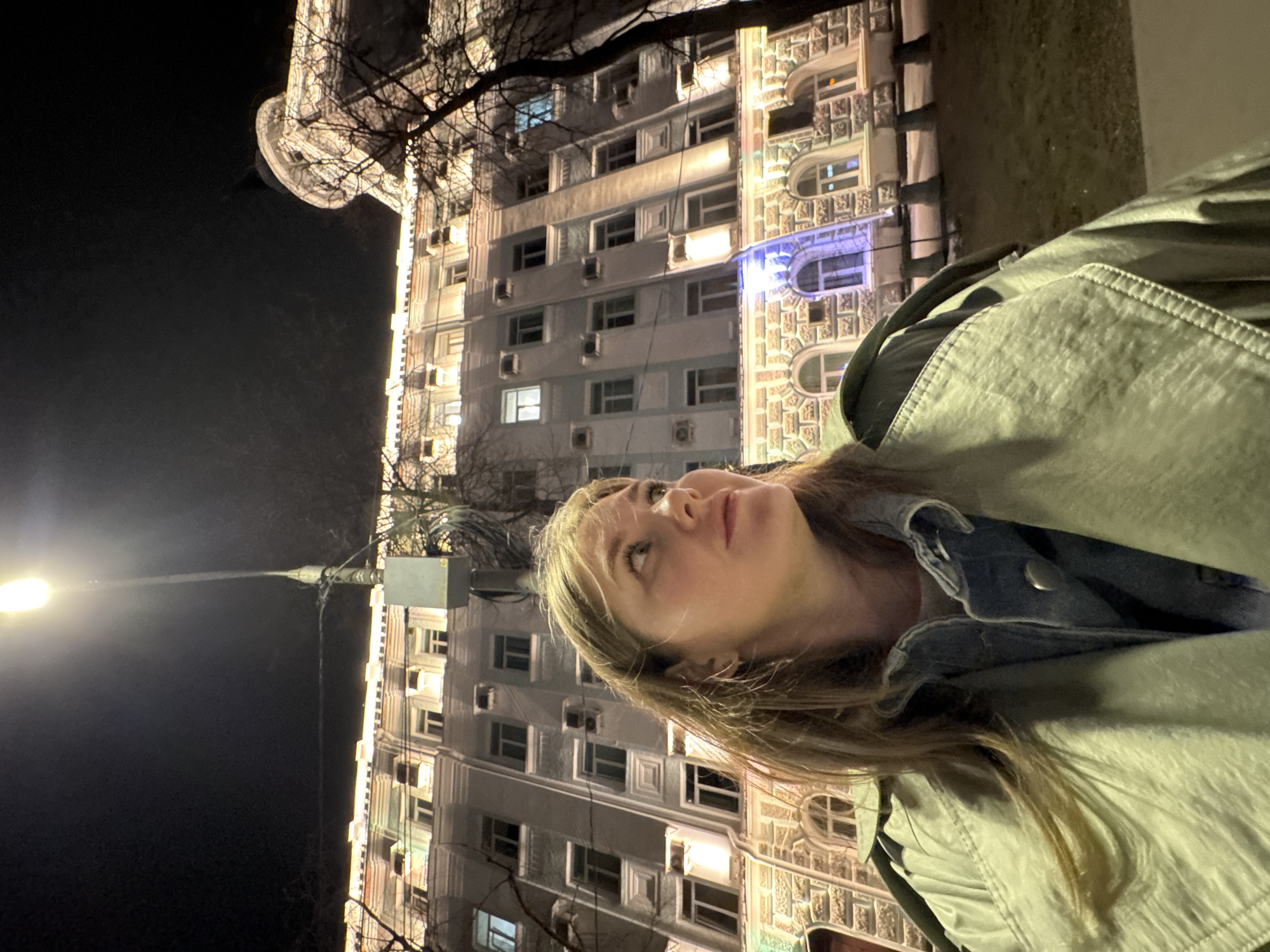 A person with blonde hair and a light green jacket is looking upwards in an image taken at night. The building in the background appears to be a multi-story structure with illuminated windows and distinct architectural details, including decorative elements and air conditioning units affixed to the exterior. To the left, a streetlight is visible, casting a bright glow. The building's design and ornamentation suggest an urban setting. Based on these elements and the location information provided, the image likely depicts a scene in Perm, Russia.