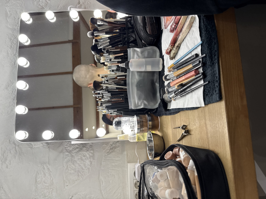 An indoor scene in Paris, France, displays a professional makeup station set up on a light-colored wooden countertop. A large rectangular vanity mirror, illuminated by ten visible circular bulbs on its frame, dominates the background. Reflected in the mirror is the back of a person's head with closely shaved or bald scalp, positioned centrally, and partially to their left, the reflection of another individual with dark hair, possibly a makeup artist. Above the reflections, a wooden beam structure is visible.
On the countertop, a diverse array of makeup tools and products are arranged. To the far left, a black transparent zippered bag contains numerous small, identical containers of liquid makeup. Adjacent to it, a small metallic bowl and a pair of silver scissors rest on the wood. Further back, a spray bottle with yellow liquid, stacked cylindrical containers of powders, and a clear plastic dispenser are visible. In the mid-ground, two black compartmentalized holders overflow with a dense collection of makeup brushes of various sizes and types. To the right, a clear plastic squeeze bottle with a pointed nozzle stands upright. In the foreground on the right, several used makeup brushes, some stained with reddish residue, are laid out on a white paper towel, which rests on a dark towel or cloth. The wall in the background on the left features a white textured finish, and an electrical outlet with two circular sockets and a grounding pin is mounted on it.