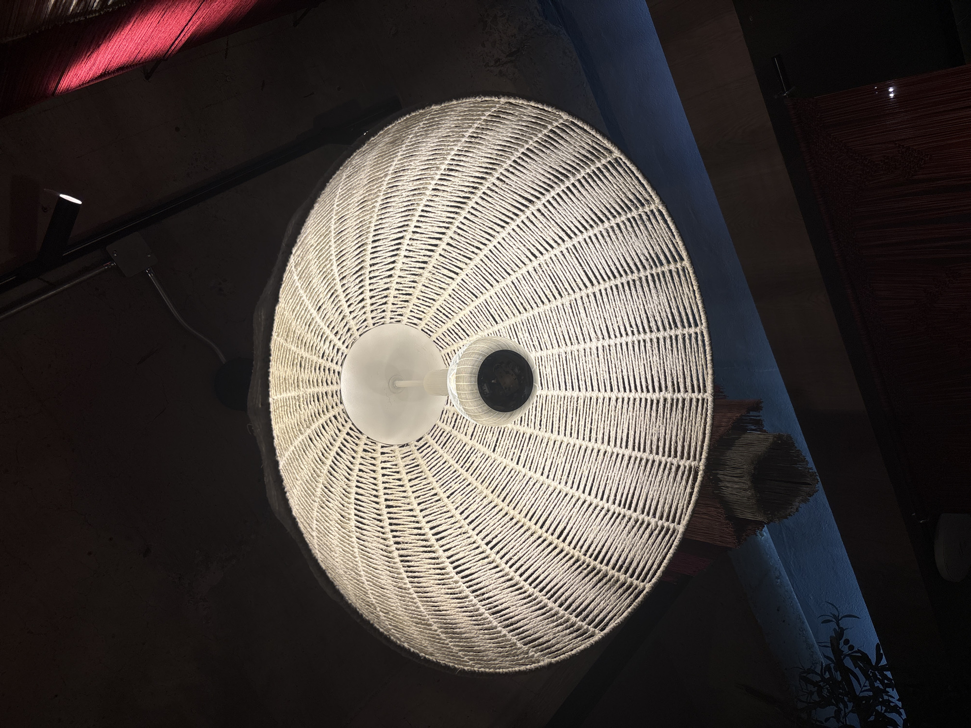 A low-angle shot reveals a woven pendant lamp illuminating an interior space. The lamp, circular and composed of interlaced white material, features a frosted white diffuser shielding a bulb. The environment is dimly lit, with a red hue cast in the upper left corner. A structural beam runs diagonally across the scene, with additional fixtures and elements visible on the edges. In the bottom right corner, plants are silhouetted against a blue wall. It is inferred that the location is in Mexico City, Mexico.