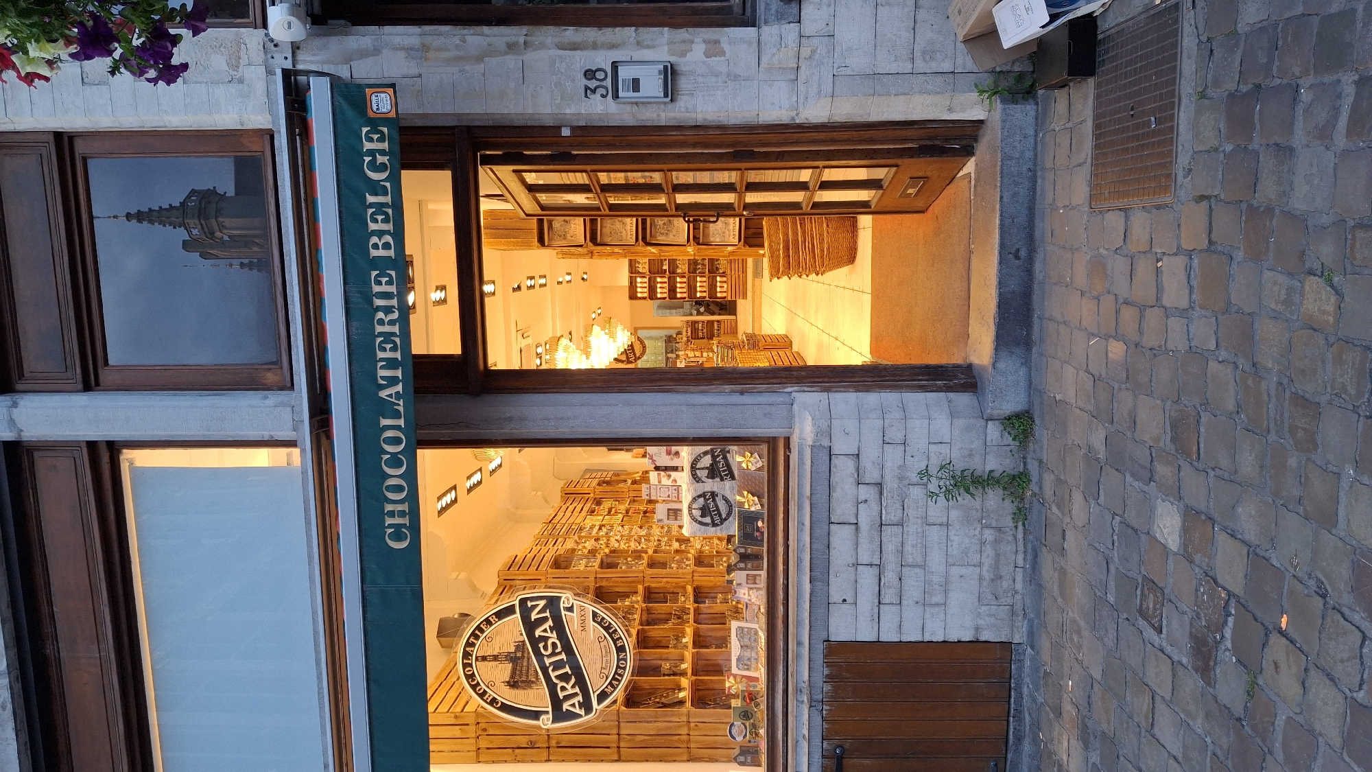 The image shows the exterior of a Belgian chocolaterie in Unknown, Unknown. The facade features a stone wall and a large display window filled with wooden shelves of chocolates. Above the window, an awning reads "CHOCOLATERIE BELGE". The entrance to the shop is an open wooden door revealing a brightly lit interior with more chocolate displays and chandeliers. A house number, "38", is visible on the exterior wall next to the door. The cobblestone street in front of the shop is wet, suggesting recent rain. A few plants are growing in cracks in the wall. There are no people visible in the scene.