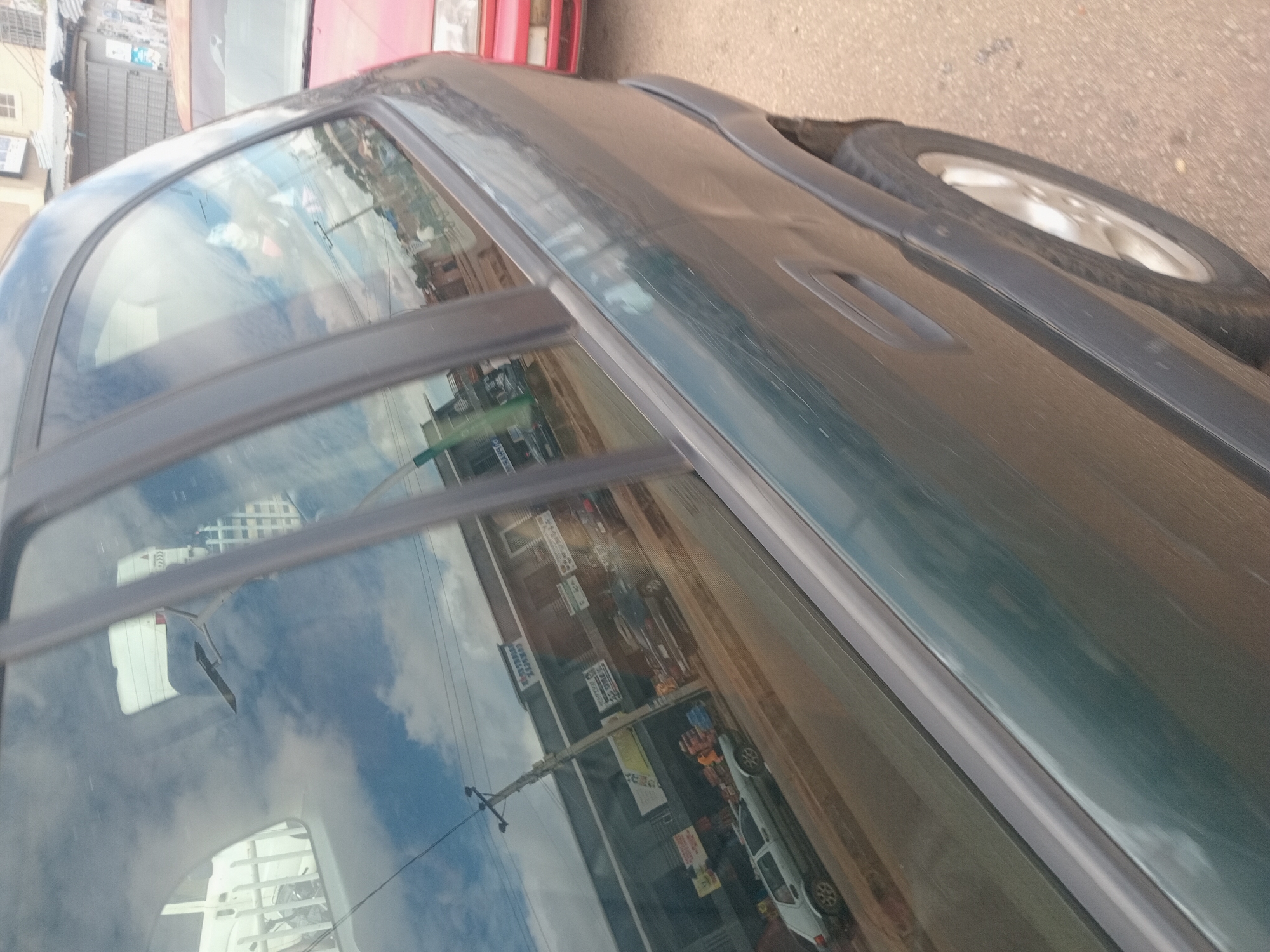 The image is a side view of a dark green car parked on a street in Bukuru, Nigeria. The car's windows reflect the sky, clouds, buildings, and a street scene. A red car is parked to the right of the green car. The street appears to be paved with asphalt and dirt. Buildings visible in the reflection include shops with signage and what appear to be residential structures. A utility pole with wires is also reflected in the window. There are no people clearly visible in the immediate vicinity of the cars, but there may be figures in the background reflections. The overall impression is that of a typical urban street scene.