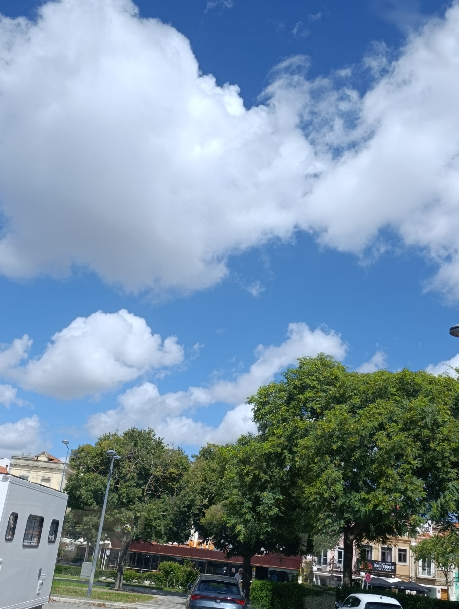 The image captures a street scene in Setúbal, Portugal, under a bright blue sky with scattered white clouds. In the foreground on the left, a white trailer or mobile unit with several windows is parked. A portion of a dark-colored car is visible to the right of the trailer. Dominating the middle ground are large, lush green trees, providing a canopy over a street. Behind the trees, buildings with a mix of architectural styles are visible, including one with a sign that reads "RESTAURANTE DOM PEPPONE PIZZARIA". A street lamp with a modern design is positioned between the trailer and the trees. The overall impression is a sunny day in an urban environment, with a clear view of the sky and the natural element of trees contributing to the setting.