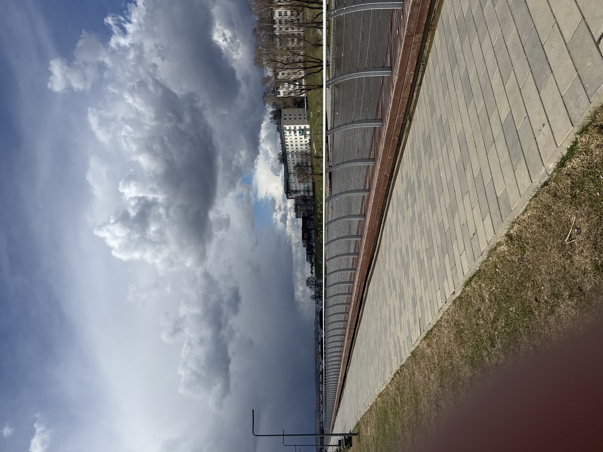 An outdoor scene in Votkinsk, Russia, captured during the daytime under a dramatic, cloudy sky. The image is composed with a paved walkway on the right, a covered structure in the middle, and buildings with trees on the left.
The foreground on the right features a light-colored paved pathway, possibly made of concrete or stone pavers arranged in a stretcher bond pattern. To the left of the walkway, there's a strip of dry, patchy grass, indicating either late winter or early spring.
Running parallel to the walkway is a long, modern-looking covered structure, possibly a promenade or seating area, with a series of arched metal supports and a translucent roof. Beyond this structure, a body of water is implied, leading to the far bank.
The background on the left is dominated by a cityscape featuring several multi-story residential buildings, likely apartment blocks, characteristic of Russian architecture. Many of the trees in front of and between the buildings are bare, suggesting the season is either early spring before leaf-out or late autumn after defoliation.
The sky is a prominent feature, filled with large, volumetric clouds ranging from bright white to dark grey, indicating varying thickness and possibly active weather. Patches of lighter blue sky are visible, particularly towards the top and center of the frame, suggesting the clouds are breaking or moving.
No people or specific activities are visible in the static scene. The lighting suggests an overcast day, but the presence of both dark clouds and brighter sky patches prevents a clear determination of specific time, though it is certainly daylight. There is no visible text in the image.