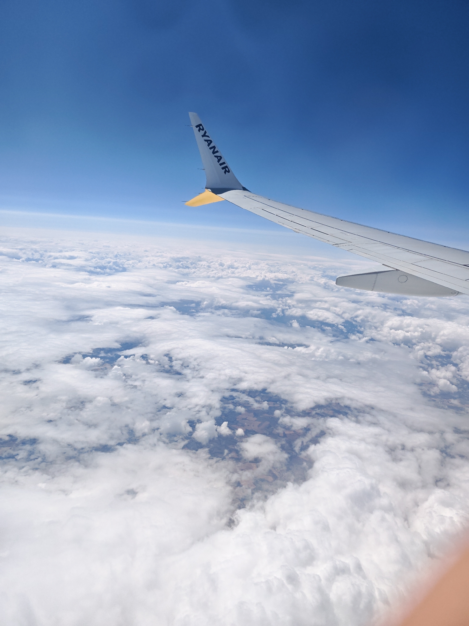 An aerial shot captures the wing of a Ryanair airplane flying above a layer of clouds. The wing, painted white with the airline's logo in black lettering, extends from the right side of the frame. A yellow wingtip is visible at the end of the wing. Beneath the clouds, patches of land can be seen, indicating a ground-level view from high altitude. The sky is a clear, bright blue. The image does not provide any specific details to identify the location as the City of Edinburgh, United Kingdom.