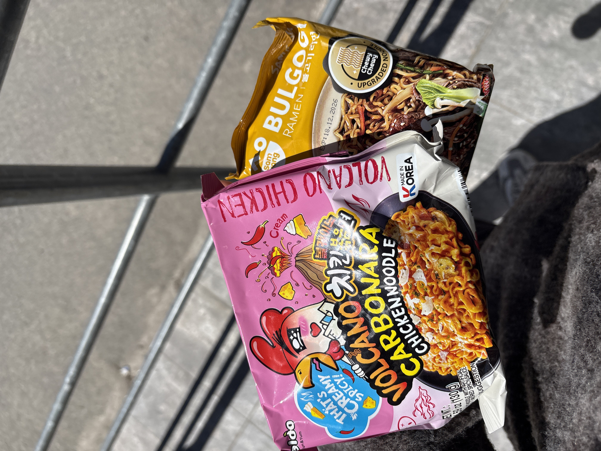 The image displays two packages of Korean instant ramen noodles, held by an unseen person whose dark, woolen clothing is visible on the right. The setting appears to be outdoors, with a concrete or paved surface and metal railings in the background, suggesting a balcony, stairs, or an outdoor urban area in Sofia, Bulgaria. Bright sunlight casts strong, crisp shadows, indicating it is daytime, likely midday or early afternoon, with clear weather.
The top package is yellow and branded "BULGOGI RAMEN" by Samyang, featuring a "Chewy Chewy UPGRADED NOODLE" label and an expiration date of "EXP:18.12.2026." The package design includes an image of ramen with beef, bok choy, and other vegetables.
Below it, a pink package of "VOLCANO CHICKEN CARBONARA CHICKEN NOODLE" is prominent. It proudly states "MADE IN KOREA" and advertises itself as "THAT'S CREAMY SPICY." The design features a playful cartoon chicken character, a volcano motif, and an image of creamy, spicy noodles. Other visible text includes "NET WT: 4.59 oz (130g)" and "NOODLE WITH ARTIFICIAL FLAVORED SPICY SAUCE." The overall activity is the display of these instant noodle products, possibly after a purchase.