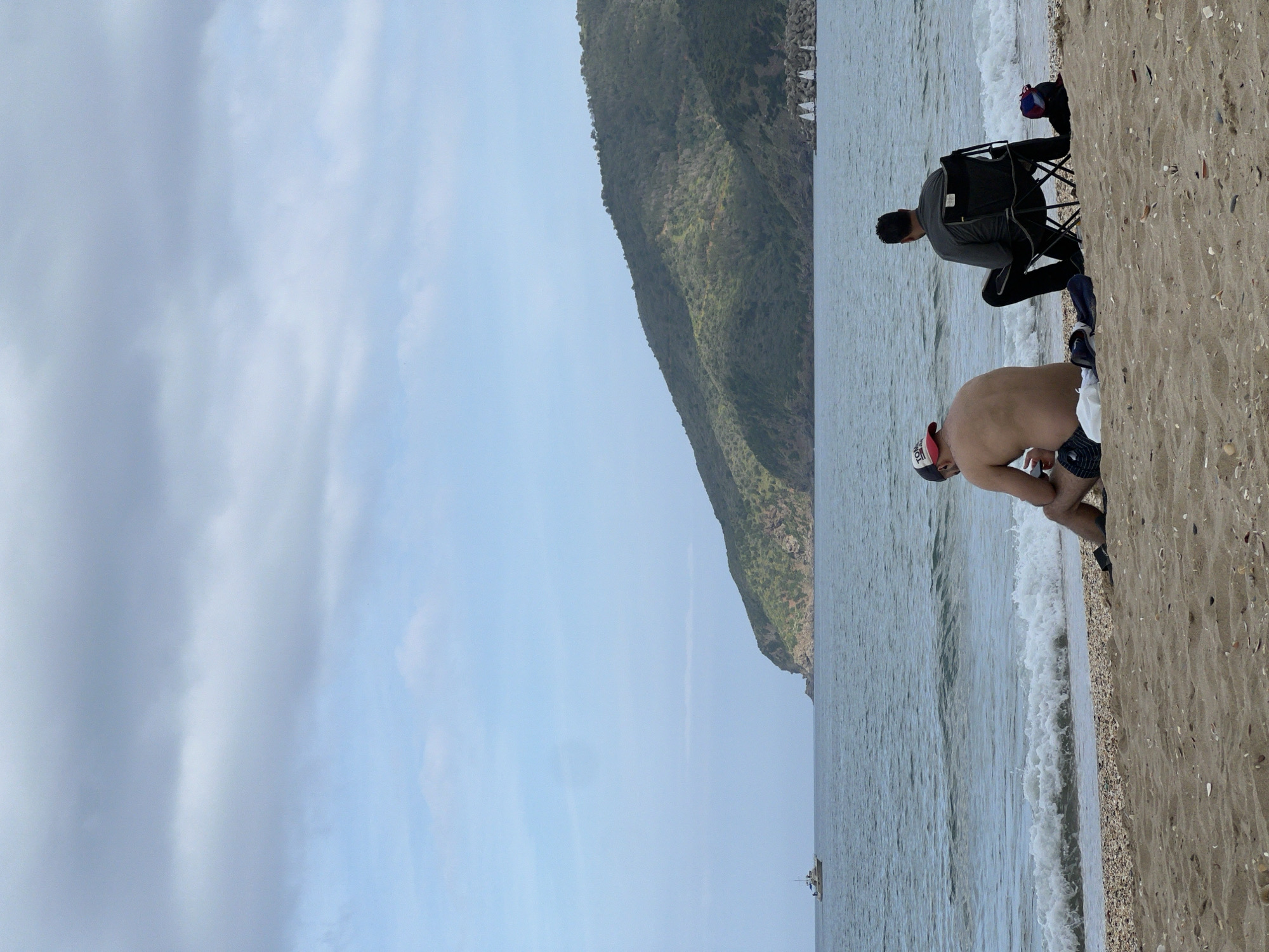 The image shows two men on a beach in M'Diq, Morocco. The first man, shirtless and wearing a red and white baseball cap and dark shorts, is bent over near the water's edge. The second man, wearing a gray long-sleeved shirt and dark pants, is sitting in a black folding chair next to a black backpack. The beach consists of sand and small pebbles. In the background, there's a hill covered in green vegetation, and beyond that, the ocean stretches to the horizon. A small boat is visible on the water in the distance. The sky is overcast.