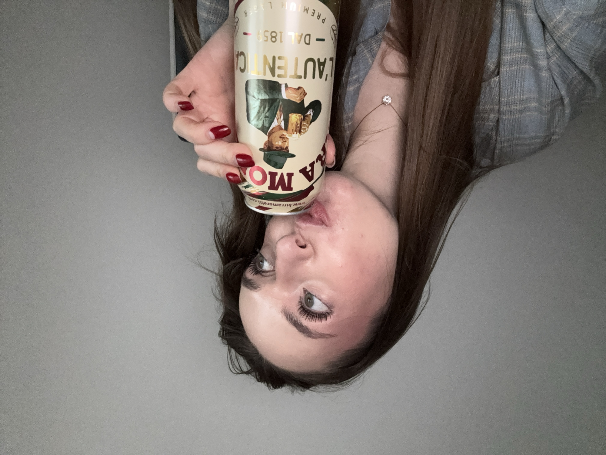 A woman with long brown hair is drinking a can of Birra Moretti L'Autentica. She is wearing a plaid shirt. The background is a plain gray wall. The location is not specified and cannot be inferred from the image.