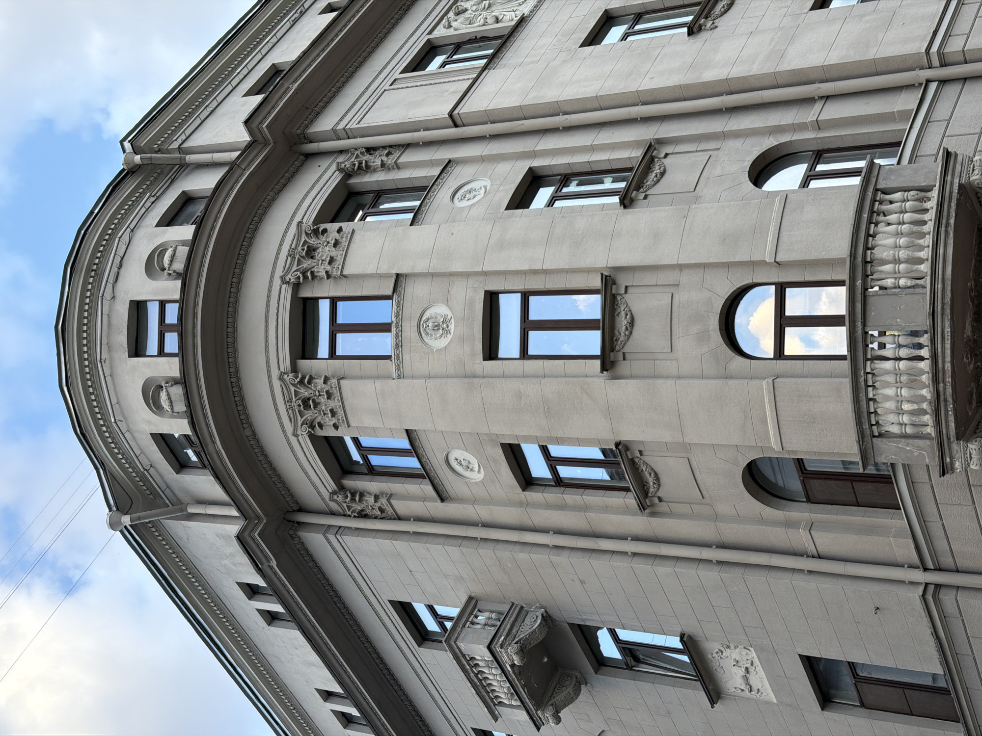 The image depicts the exterior facade of a building in Moscow, Russia. The building is constructed of gray stone and features architectural elements such as windows, balconies, and ornate carvings. The sky is visible in the upper left corner of the image.